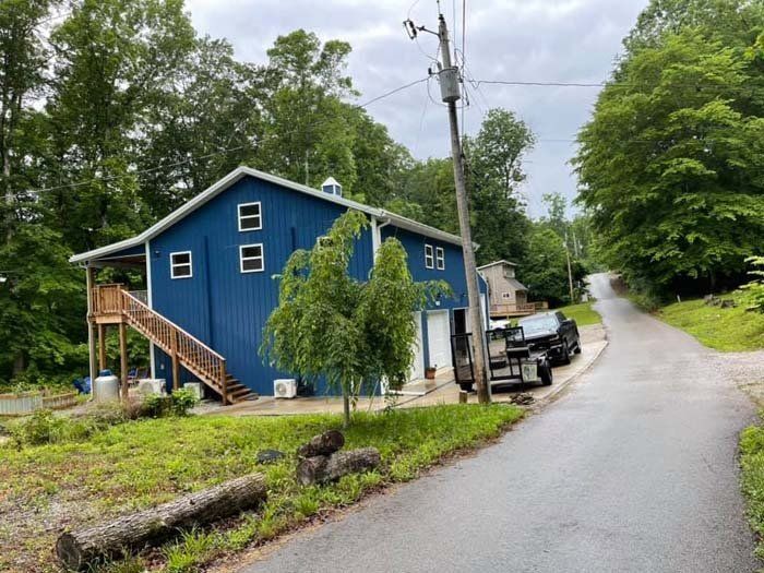 Blue barn with a wooden staircase next to a road with trees on either side. A black trailer is parked nearby.