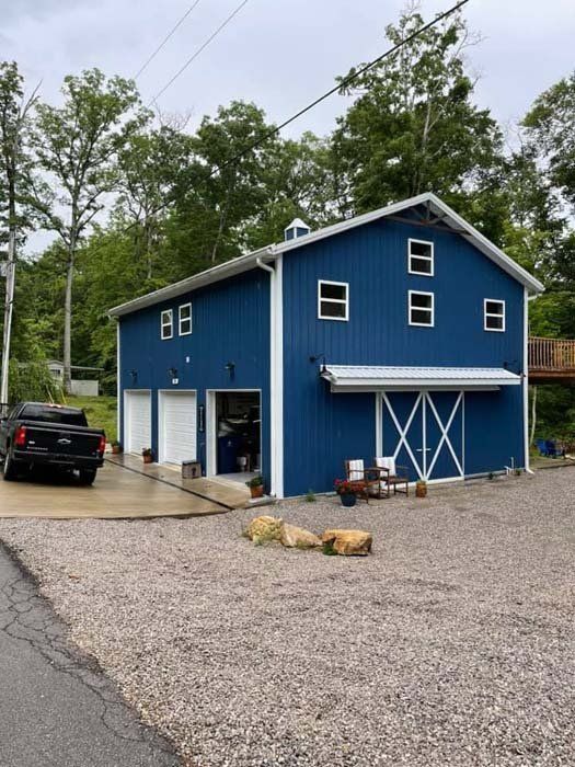 Blue barn-style building with three garage doors and a large sliding door, a pickup truck parked nearby. Gravel driveway and trees in the background.