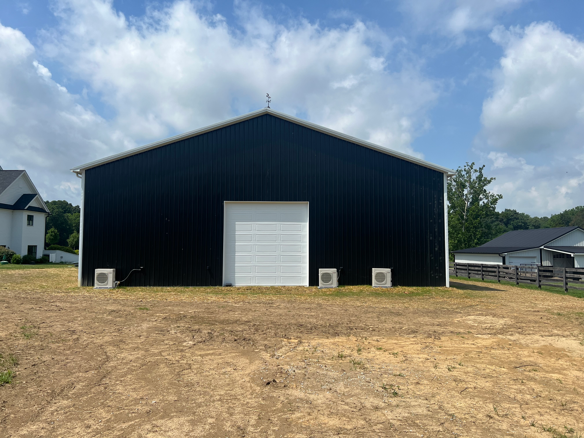 Black metal barn with white garage door, three small white boxes on the ground in front, cloudy sky overhead, and surrounding yard.