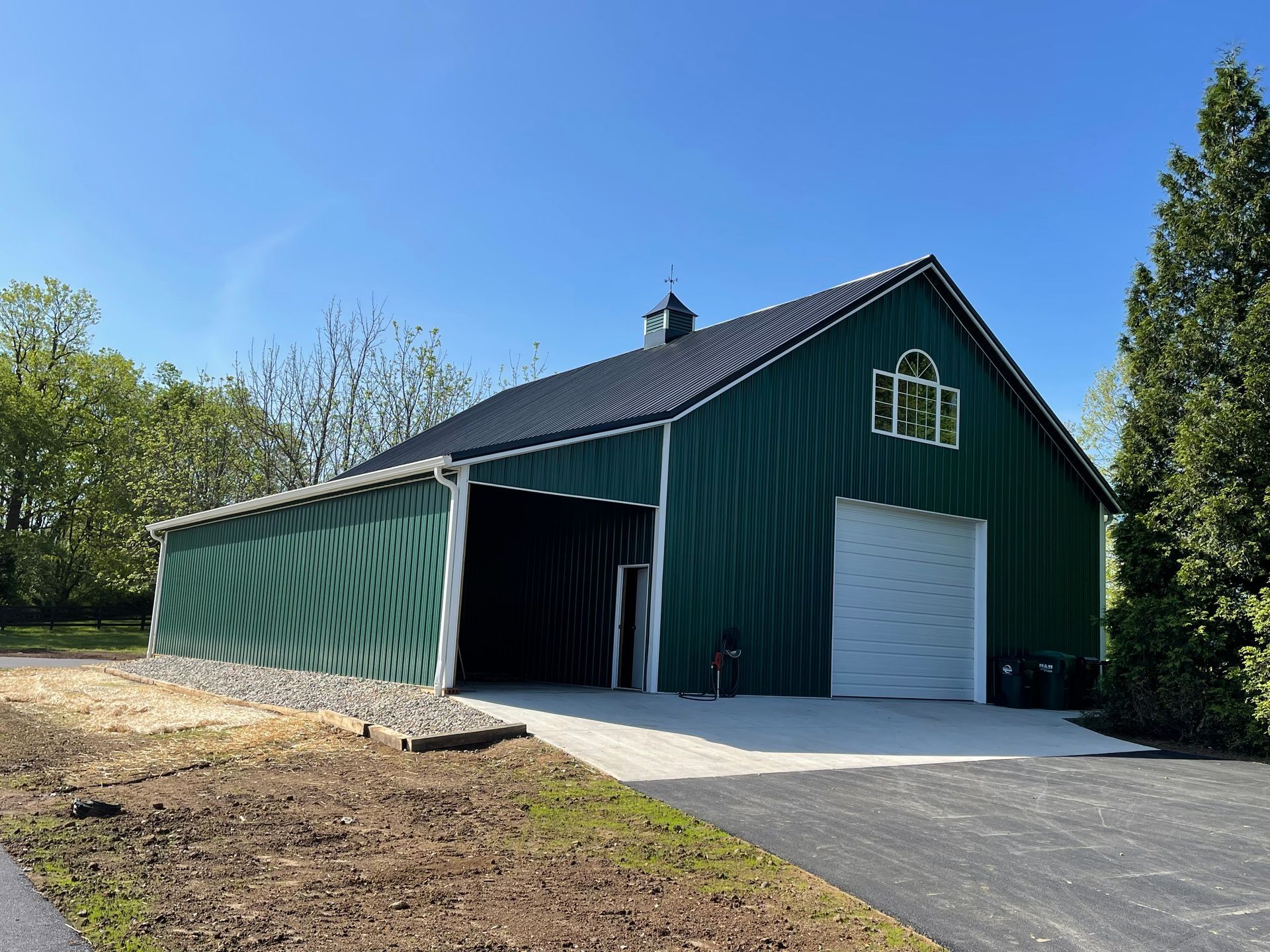 Green barn with a black roof and white garage door on a sunny day. An open-air lean-to extends from the side.