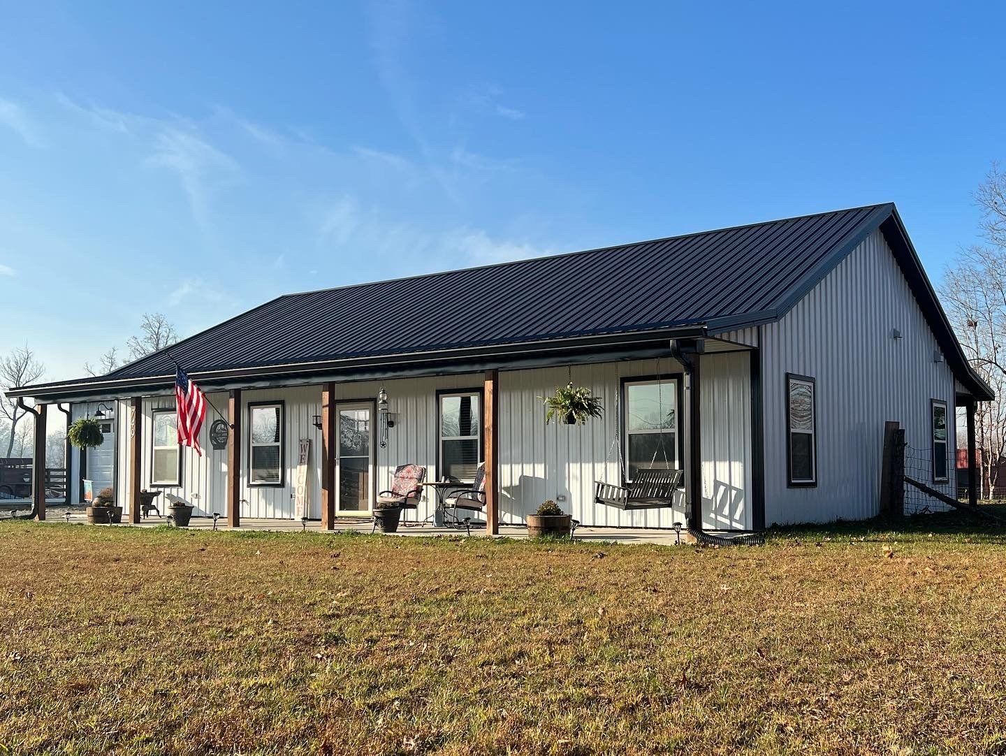 White farmhouse with a black roof and porch. An American flag hangs, and two hanging baskets are visible.