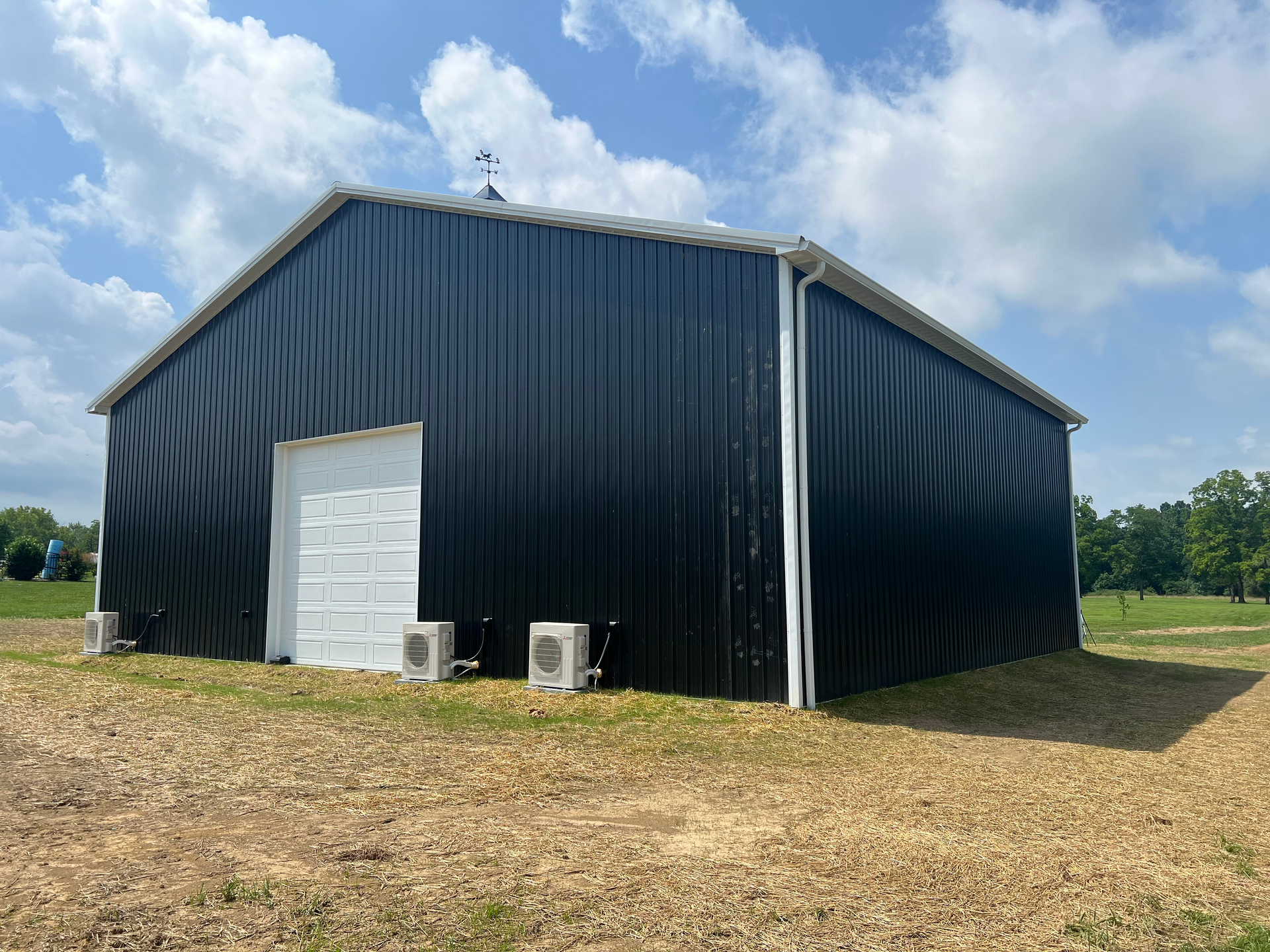 Black metal barn with white door, set in a field under a blue sky. Several HVAC units are mounted on the side.