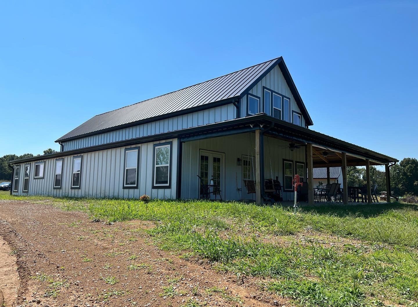 Light blue barn-style house with a black metal roof and covered porch on a sunny day. The building sits in a field.