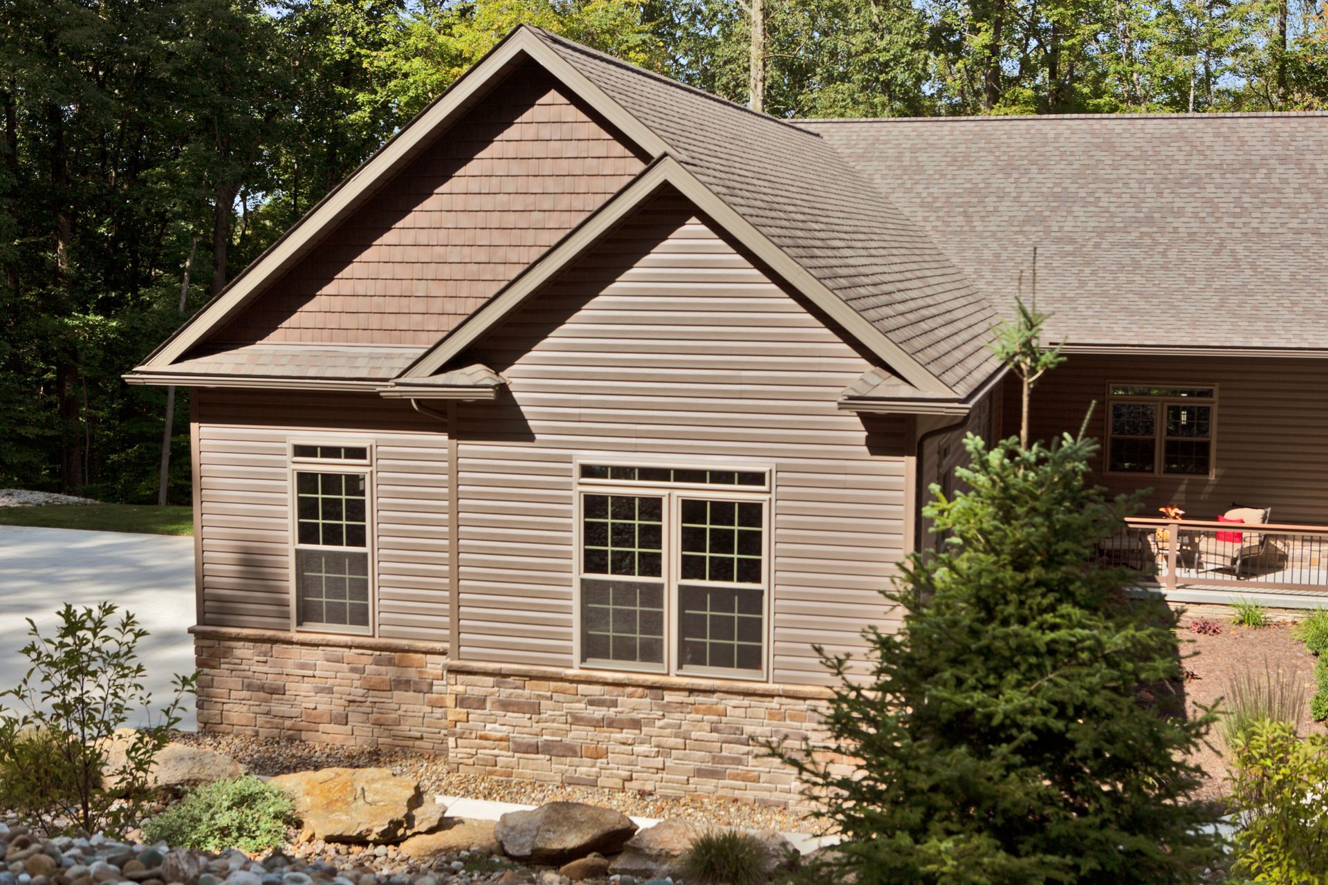 House with brown wood siding, stone base, and windows, set among trees and landscaping.