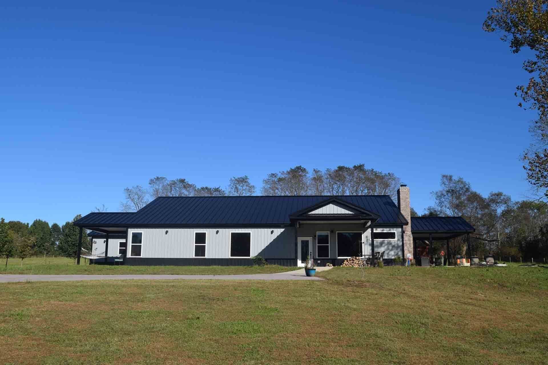 Gray house with a dark blue metal roof under a clear blue sky. A chimney rises on the right side.