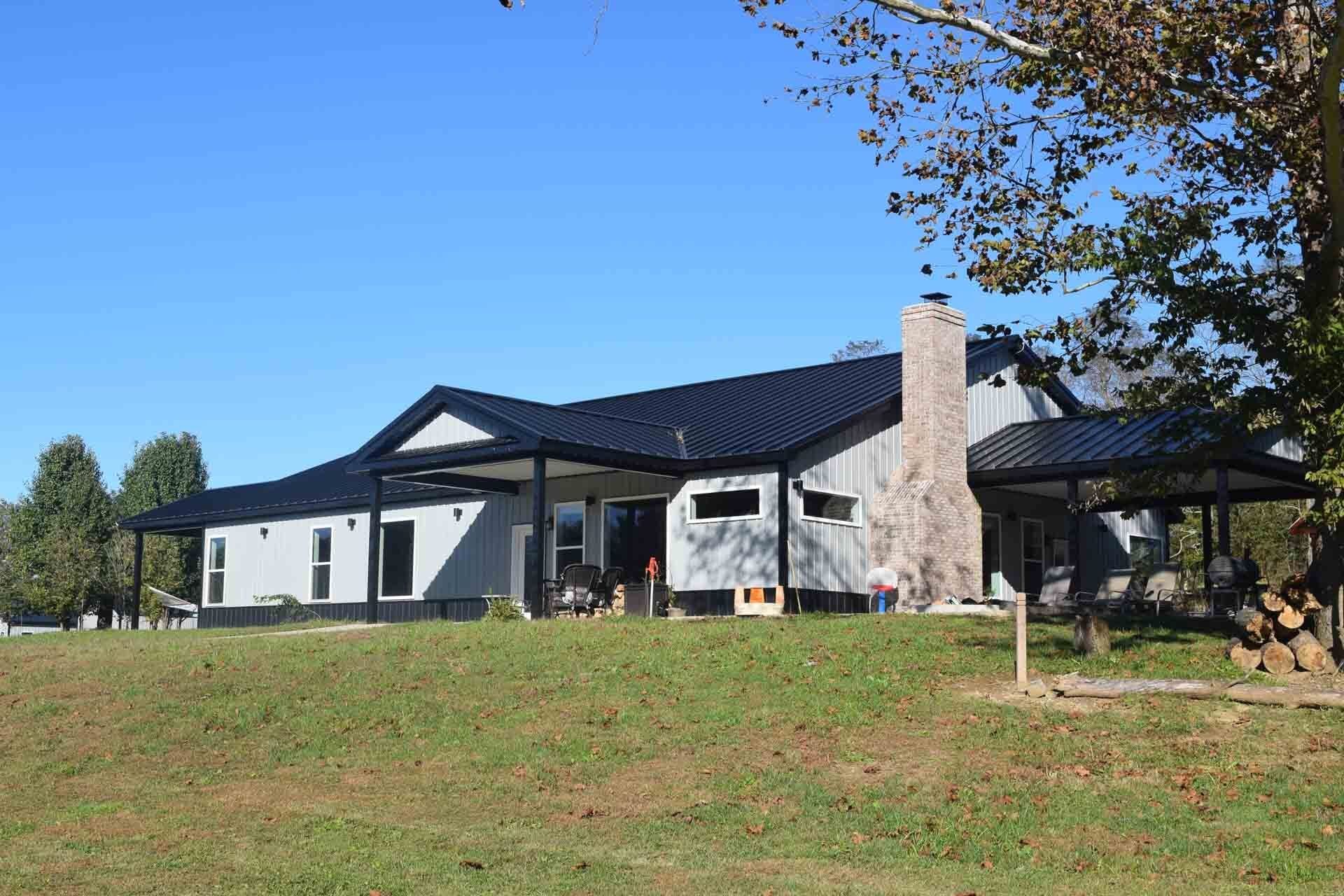 A light gray house with black roof and trim, set on a grassy hill under a blue sky. A stone chimney and firewood are present.