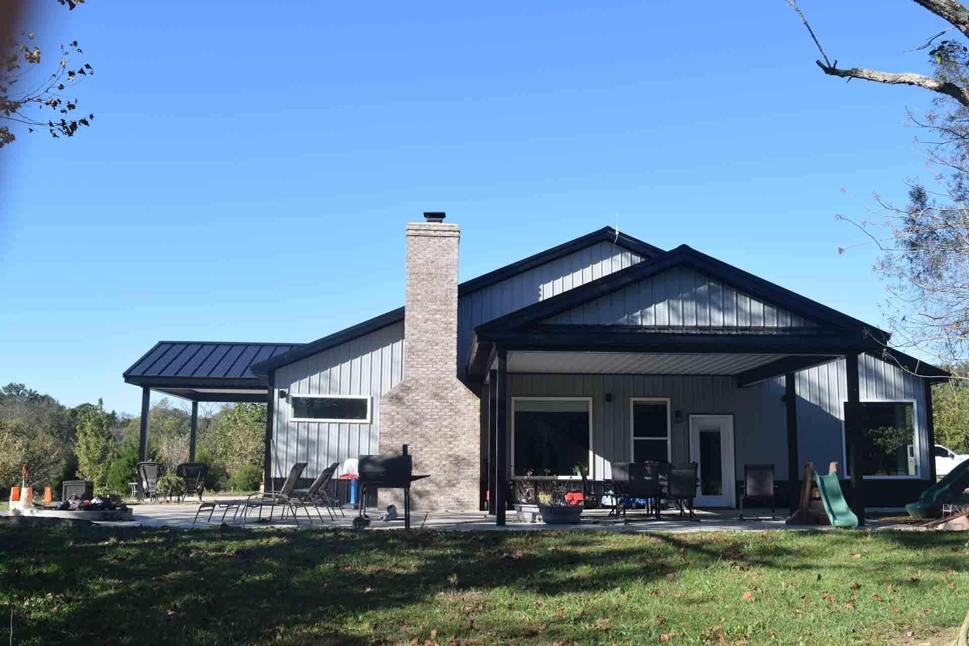 A single-story house with a brick chimney and a covered patio. The house is gray with black trim, on a lawn under a blue sky.
