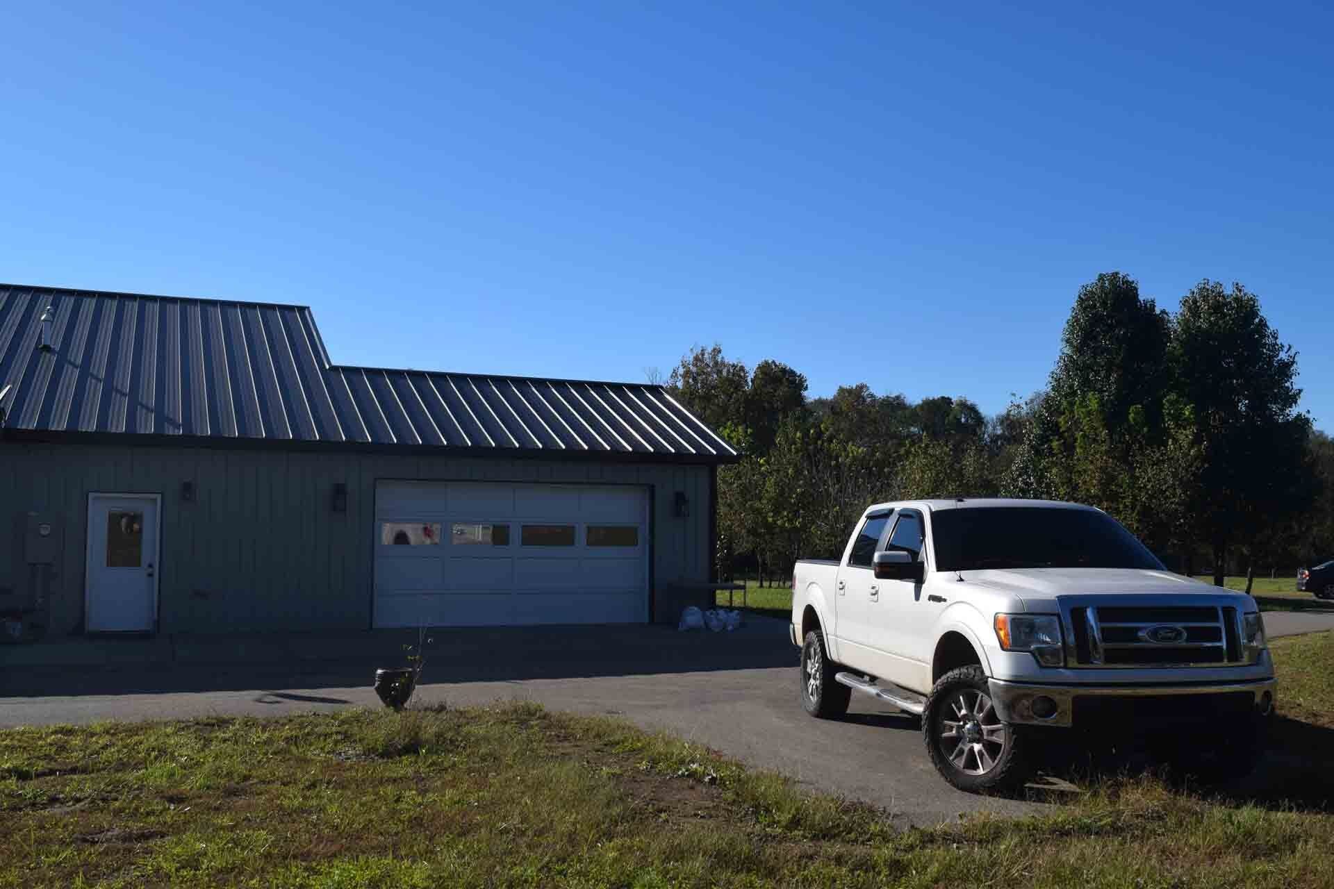 White pickup truck parked in front of a light green garage with a metal roof, on a sunny day.