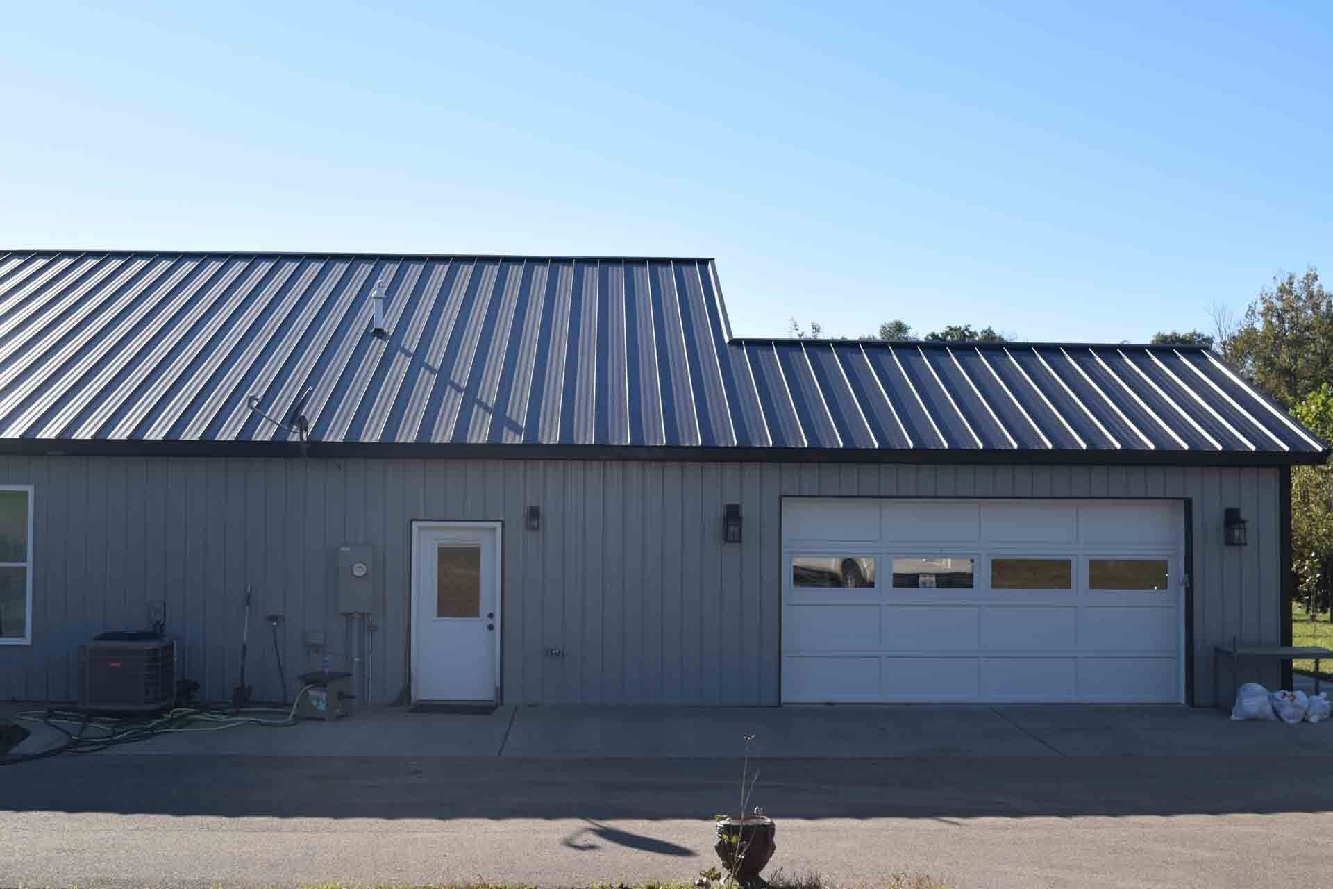 A gray building with a metal roof and white garage door sits on a paved surface under a clear sky.