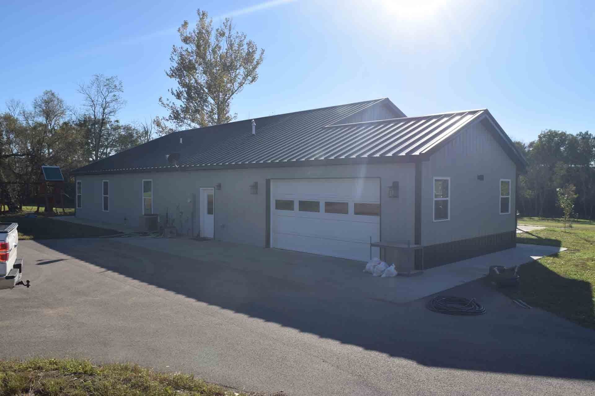 A gray metal-clad building with a garage door, set against a blue sky and surrounded by trees. A driveway leads up to it.