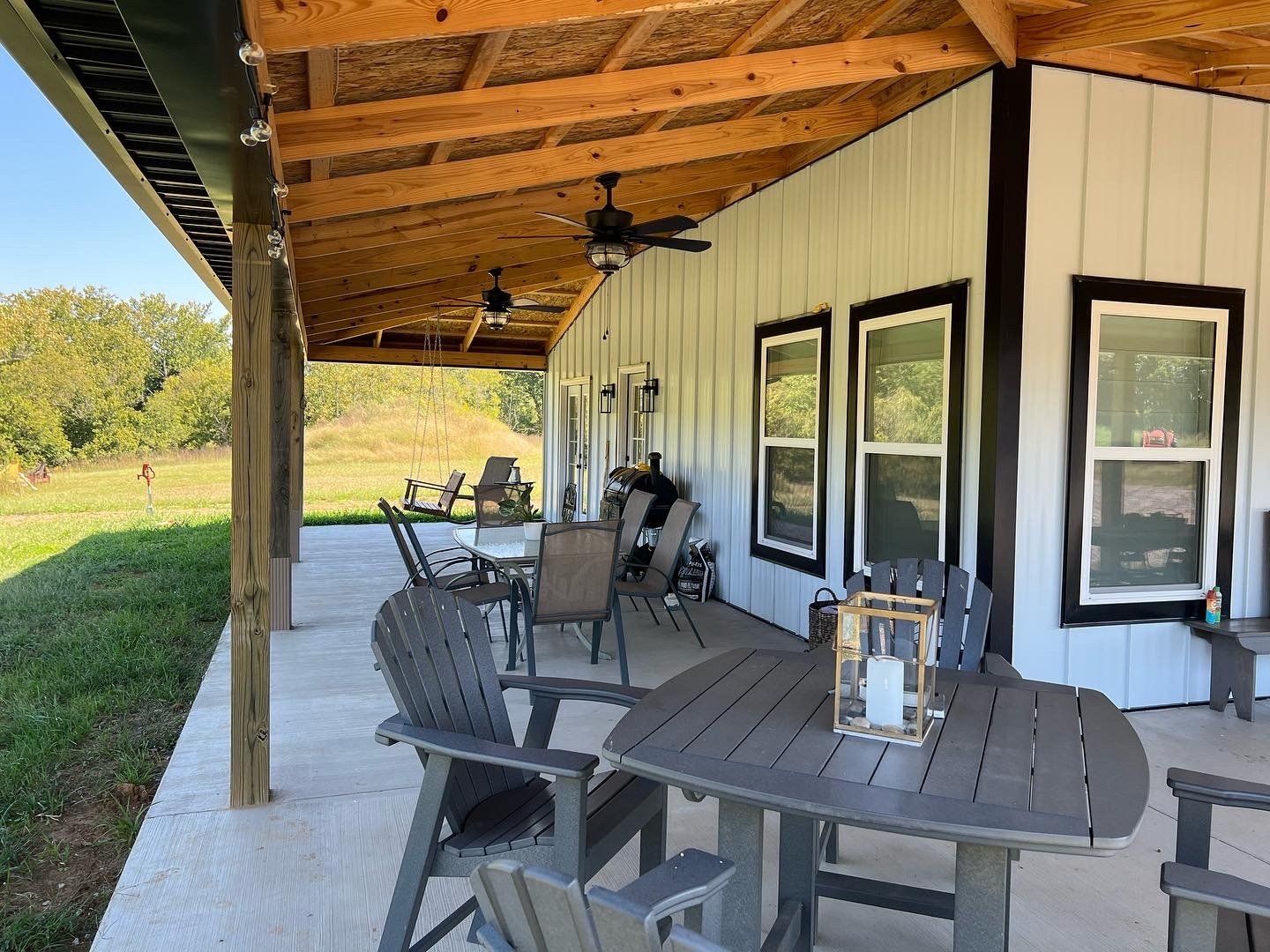 Outdoor patio with gray furniture, a dark table, and a wooden lantern. White building with black trim, overlooking a grassy landscape.