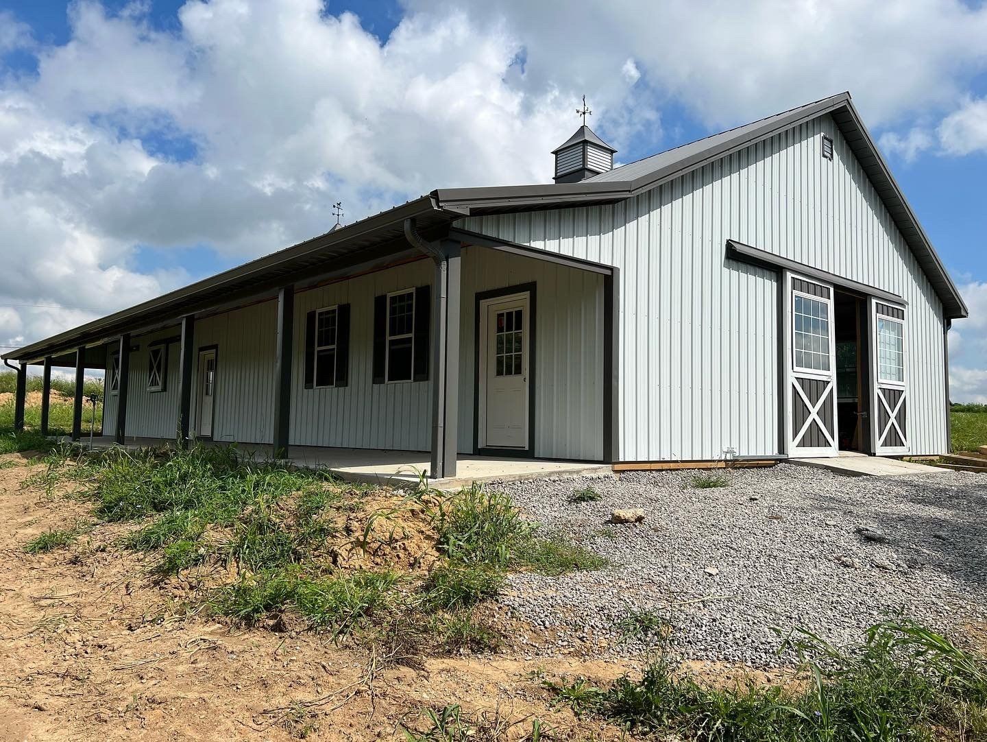 A light gray metal barn with a porch, white doors, and sliding doors. Set on a gravel foundation, under a cloudy sky.