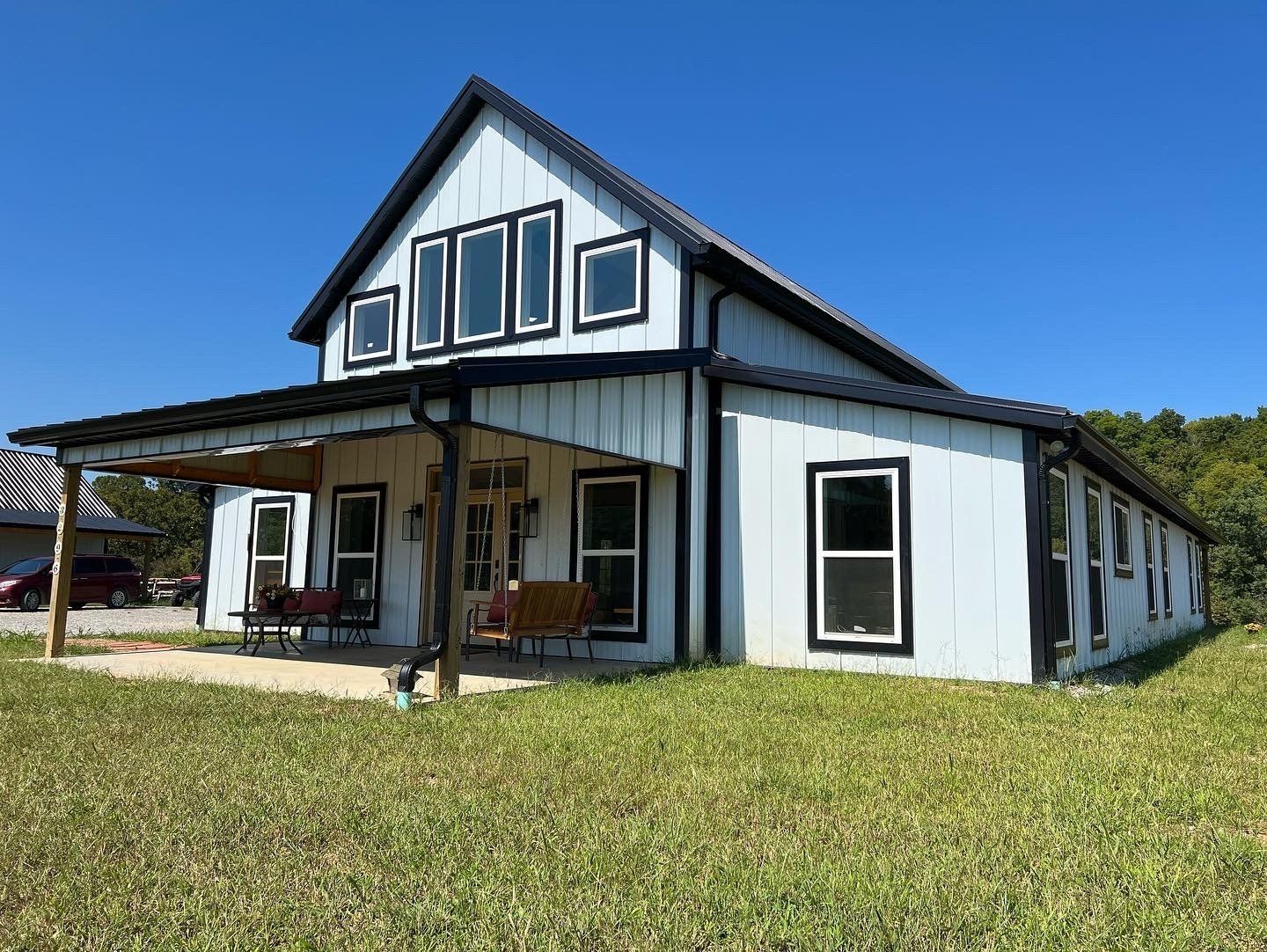 Modern white barn-style house with black trim and large windows on a sunny day. A porch with swings extends from the side.