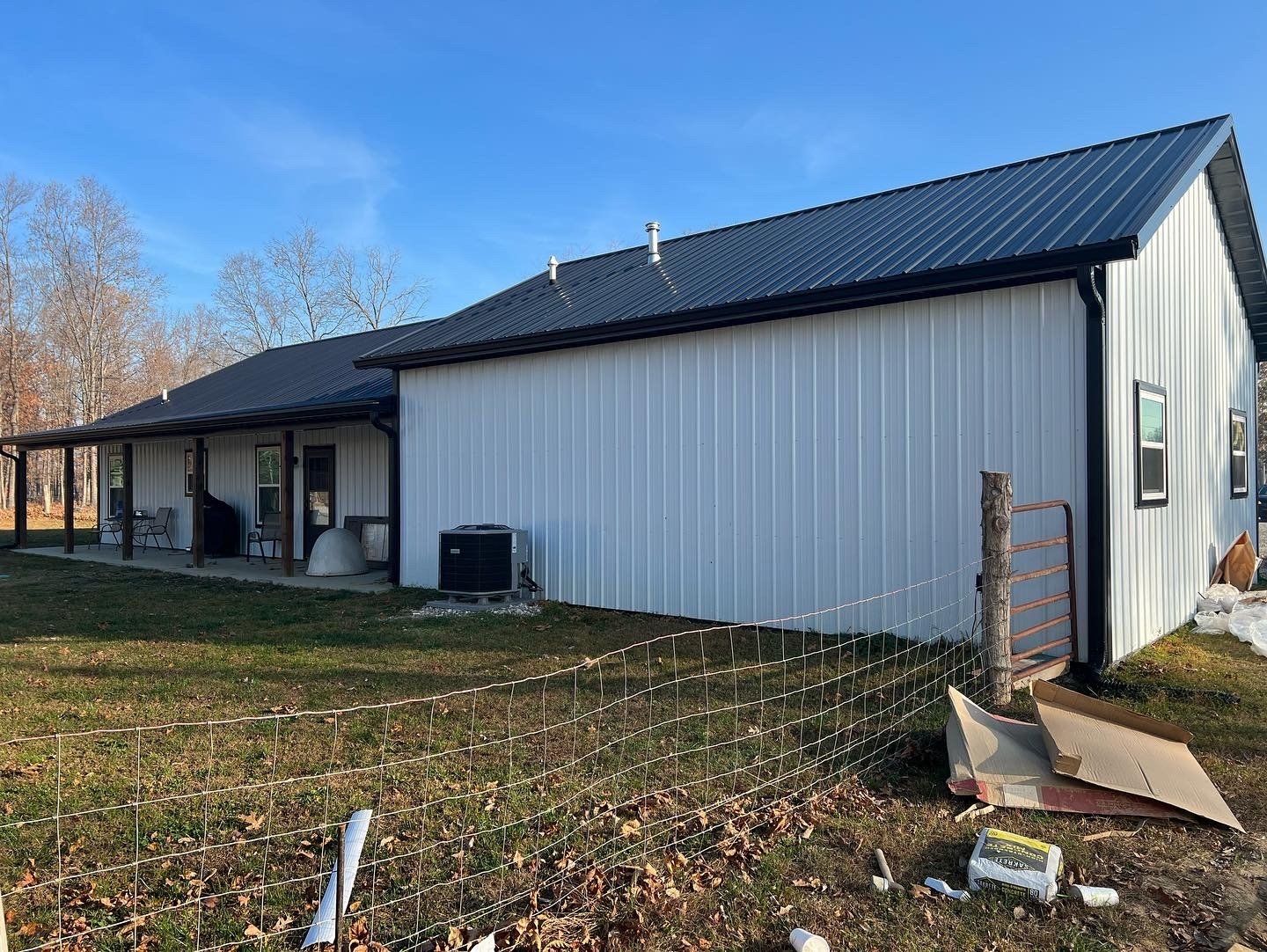 White metal-sided house with a black roof and trim. A porch extends along the side with trees in the background under a blue sky.