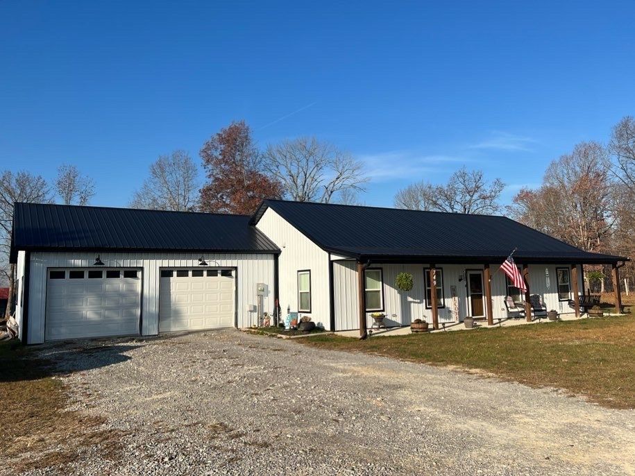 White farmhouse with a black roof, attached garage, and gravel driveway under a blue sky. An American flag hangs on the porch.