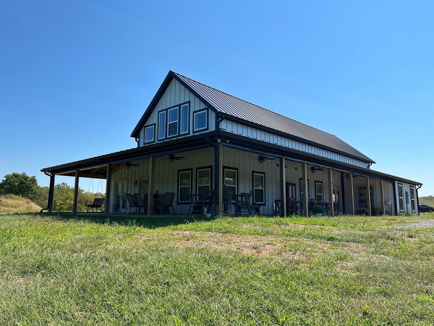 A large, light-colored barn-style house with a wraparound porch on a grassy field under a blue sky.