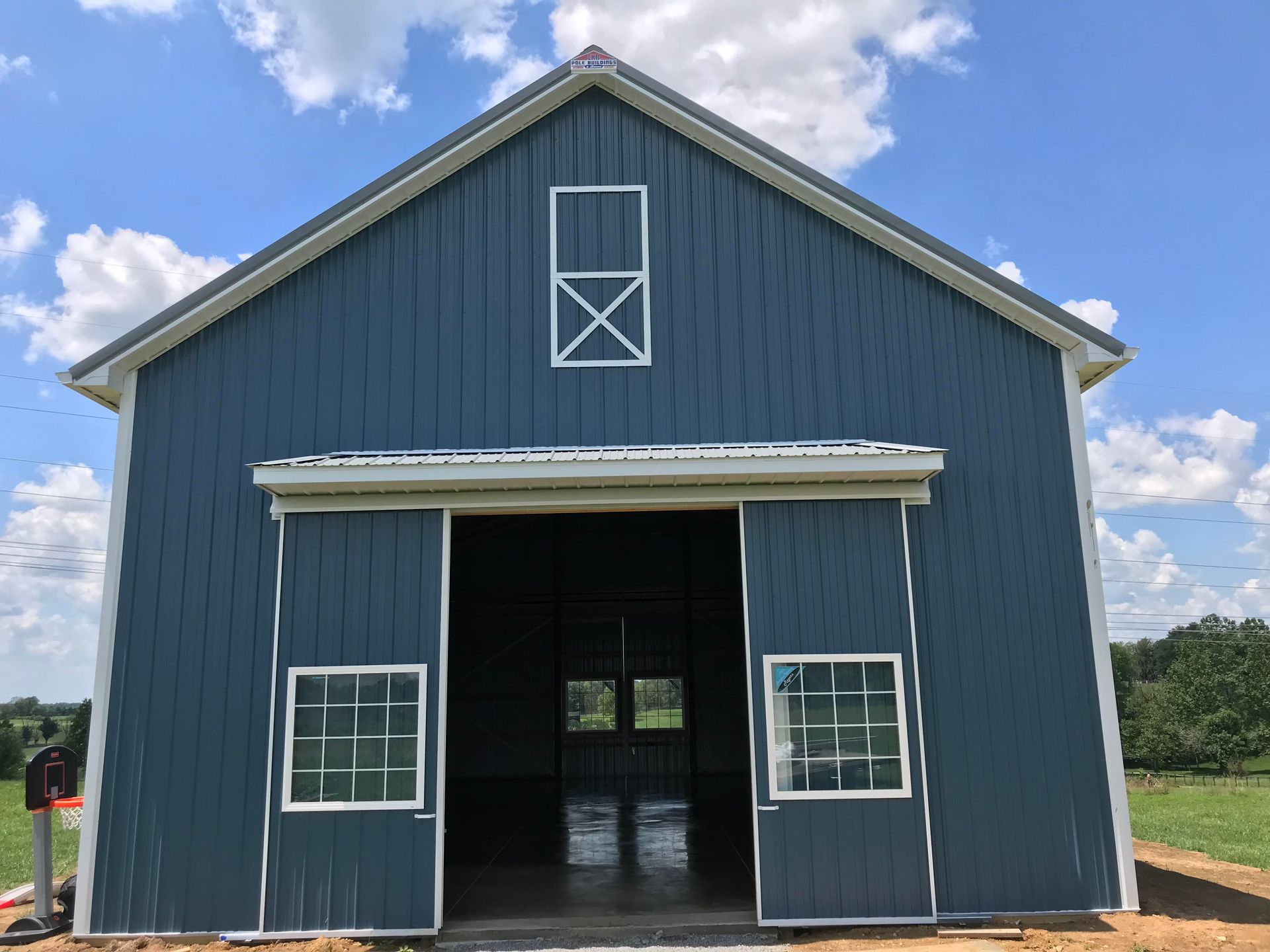 Blue barn with white trim, large sliding doors, and a window with an X design. Set in a field with a clear sky.