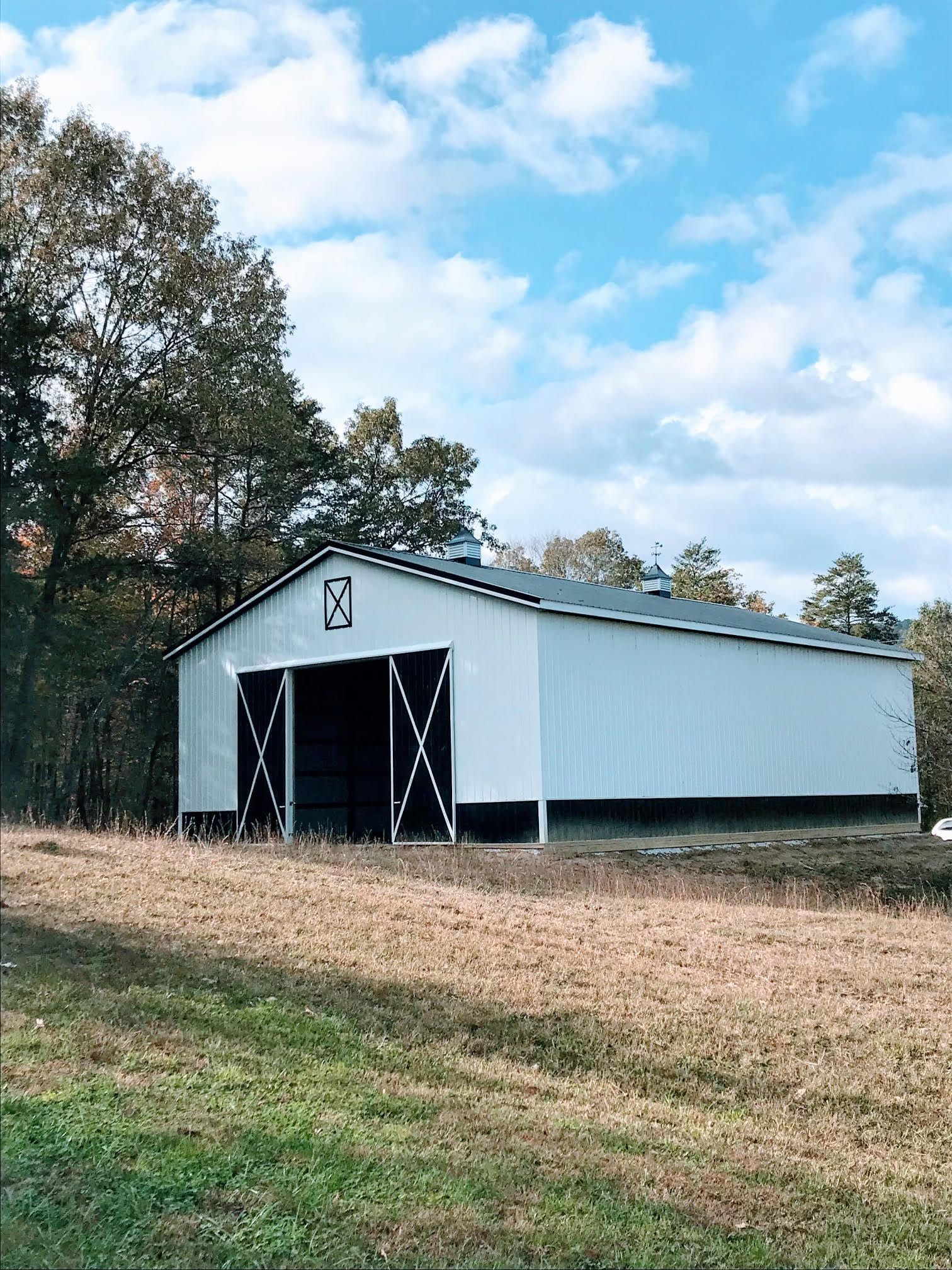 White barn with black doors sits in a field with a backdrop of trees under a partly cloudy sky.