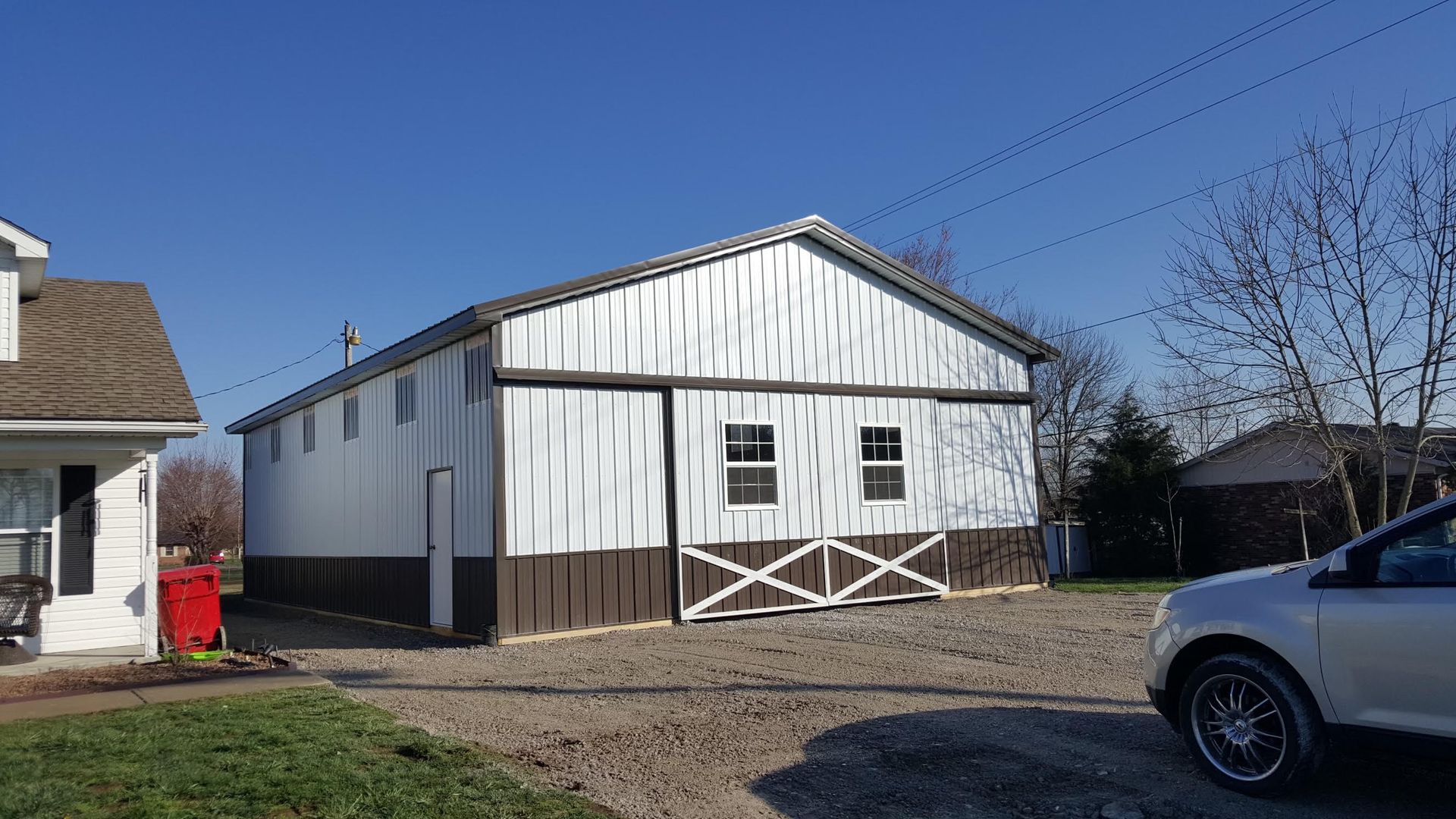 A white barn with brown trim and a sliding door, under a clear blue sky. A car and a house are nearby.