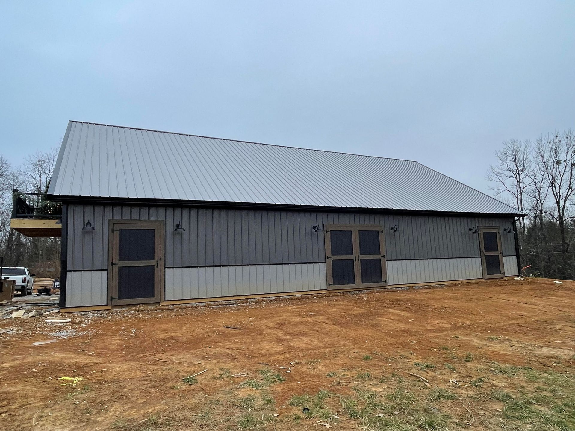 Gray metal-roofed barn with three doors, set on a slight slope, surrounded by brown earth and overcast sky.