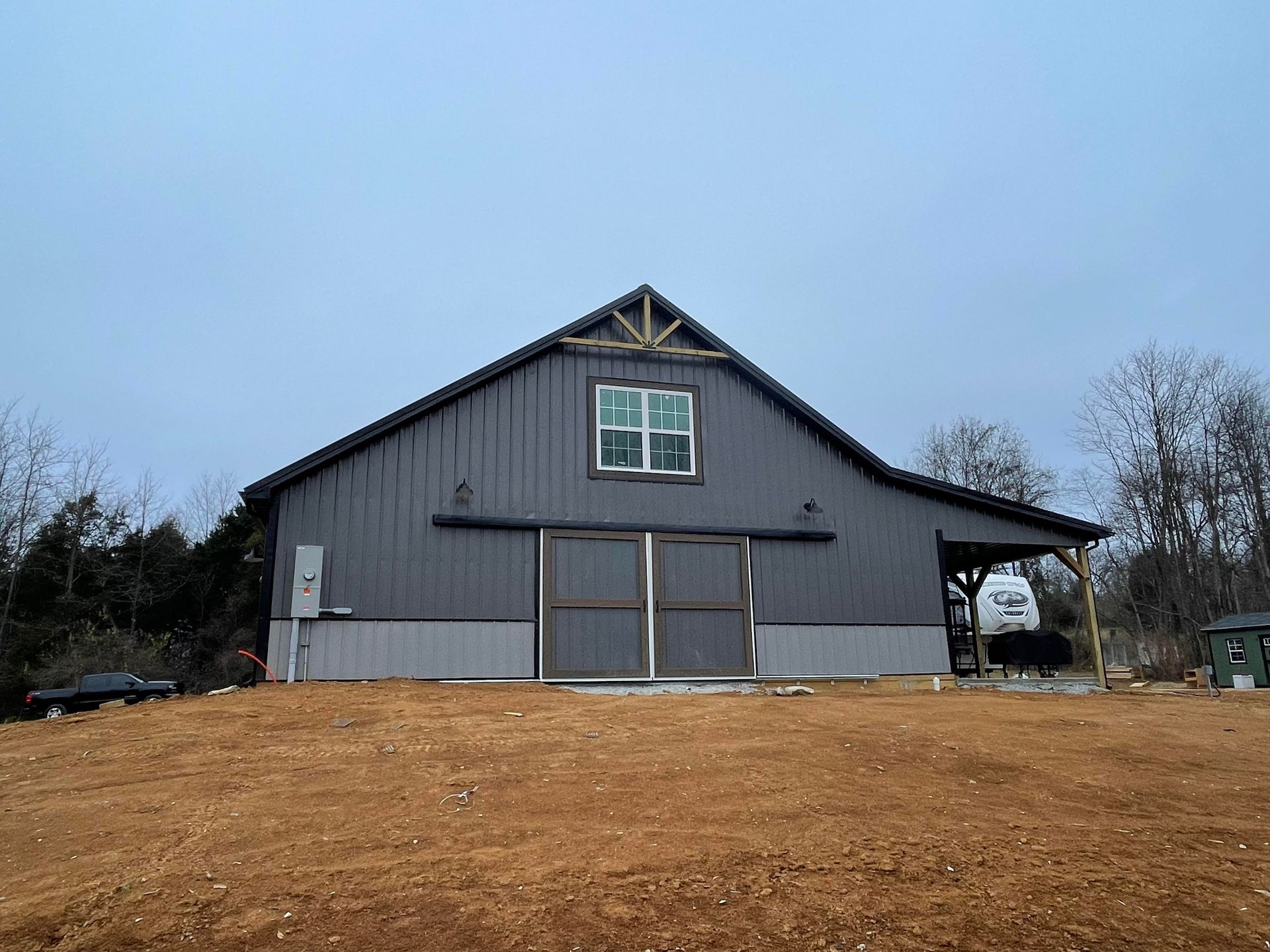Gray barn with dark gray doors and light gray lower siding against a cloudy sky. A window is in the upper center, and a propane tank sits under a small roof on the right.