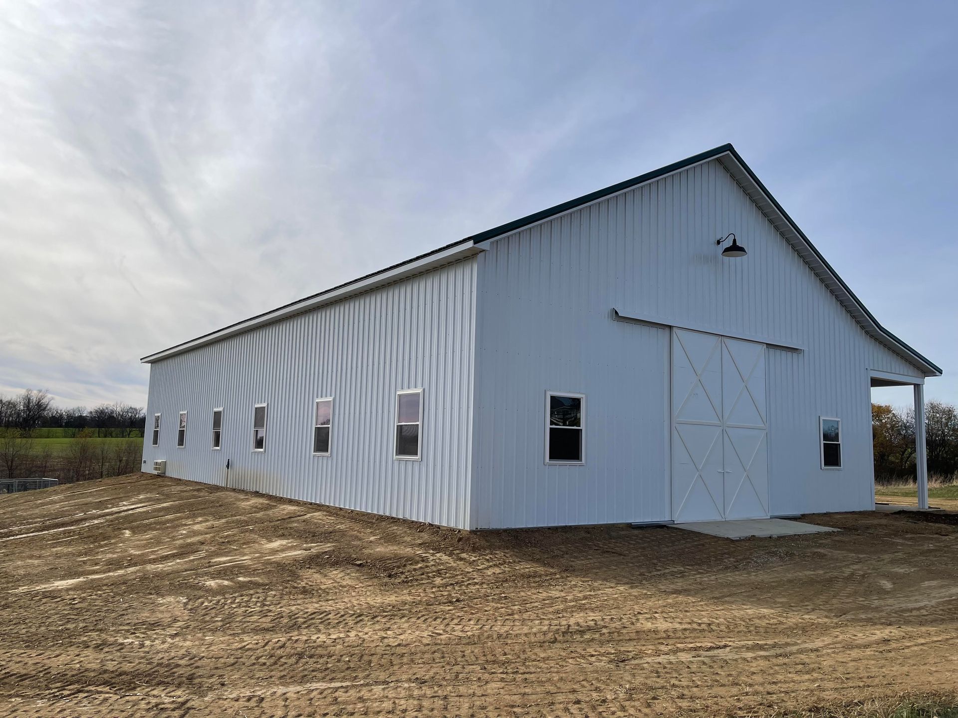 White metal barn with a black roof, set on a graded dirt lot under a partly cloudy sky.