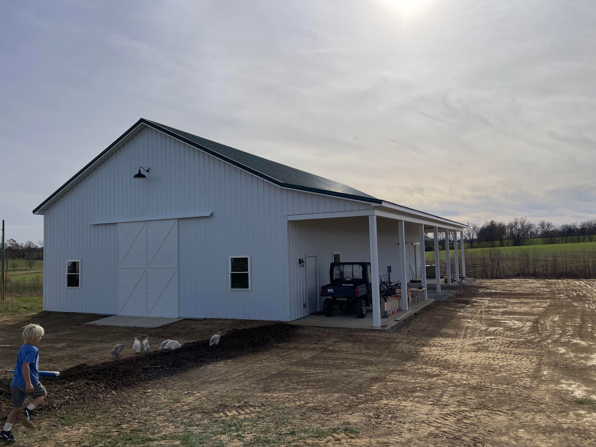 White barn with a green roof, porch, and tractor. A person is visible in the foreground; the sky is cloudy.
