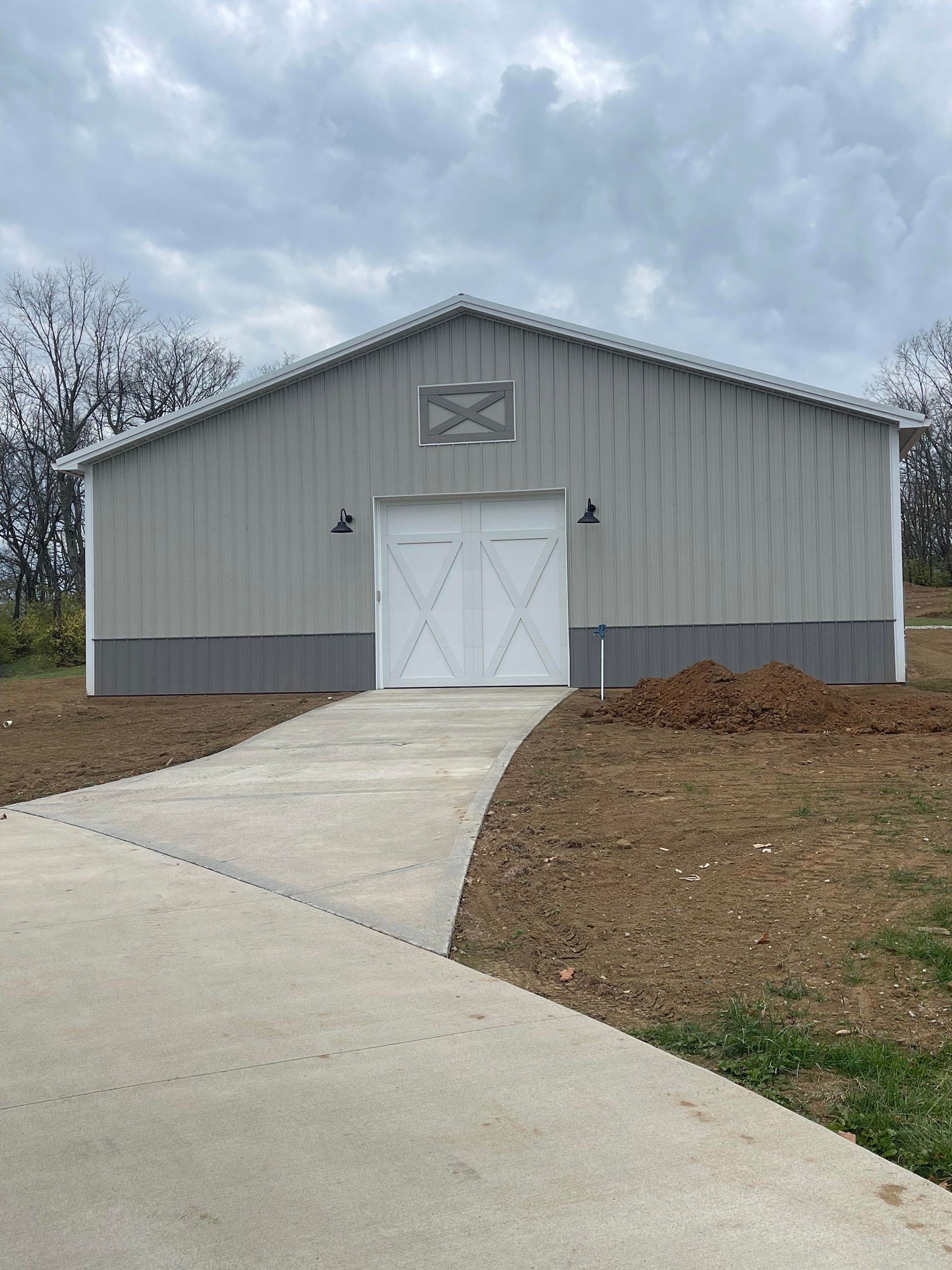 A light gray metal barn with a white door and trim, set on a concrete path against an overcast sky.