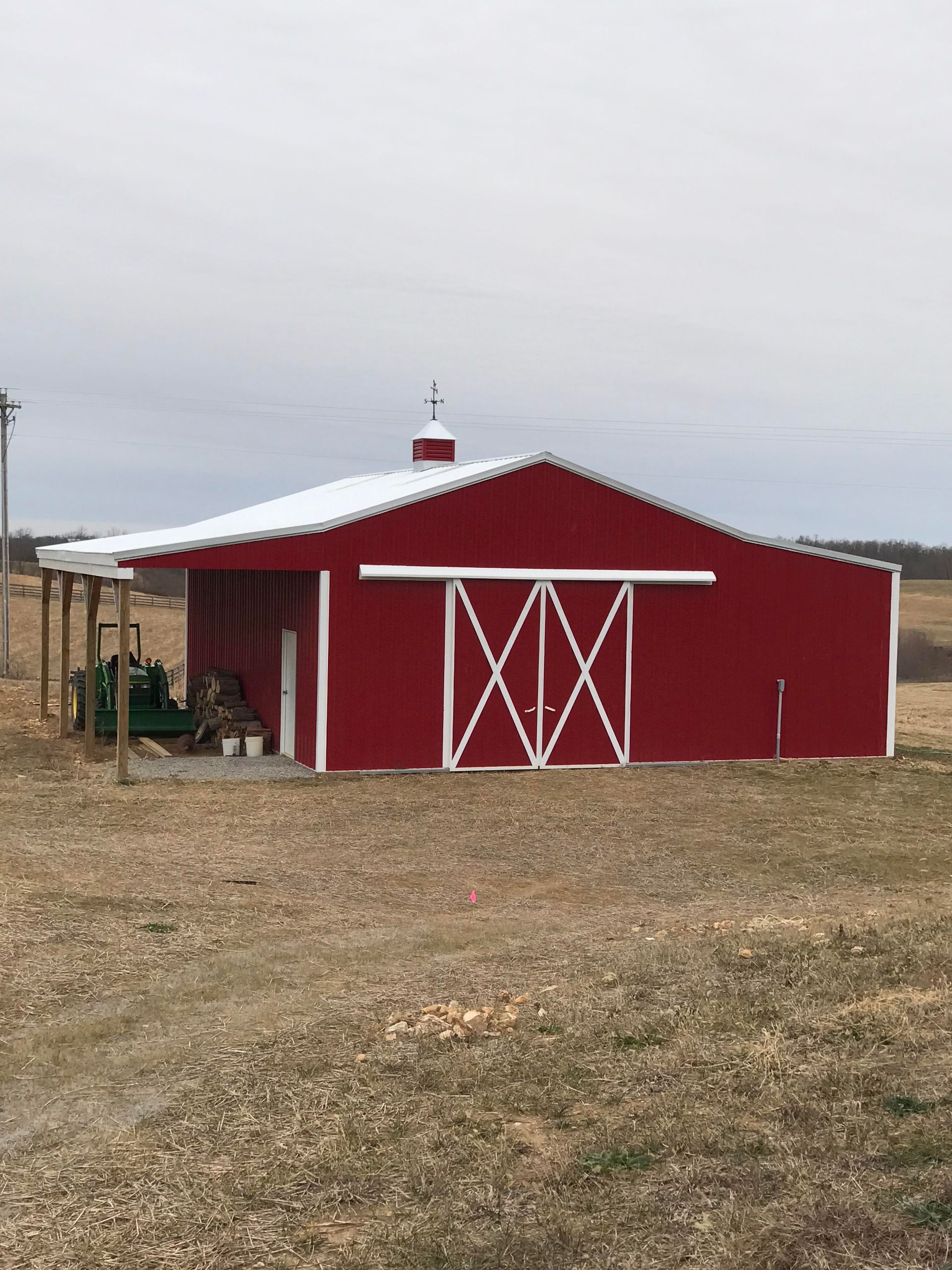 Red barn with white trim and sliding doors, set in a field under an overcast sky. A shed roof extends from the side.