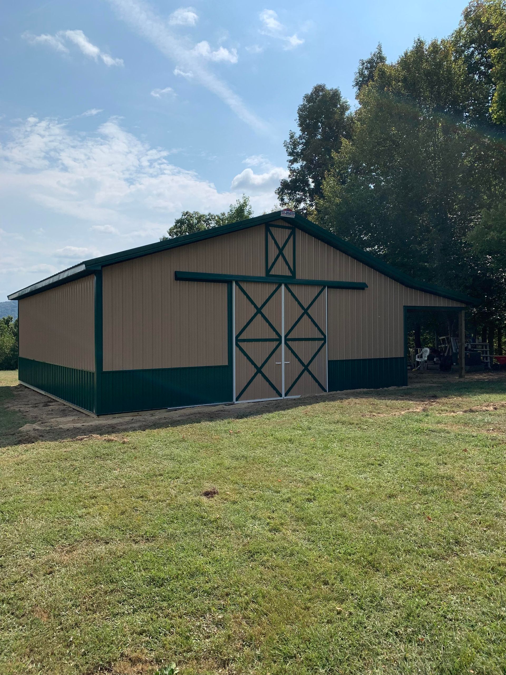 Tan and green barn with a sliding door, set in a grassy field with trees under a blue sky.