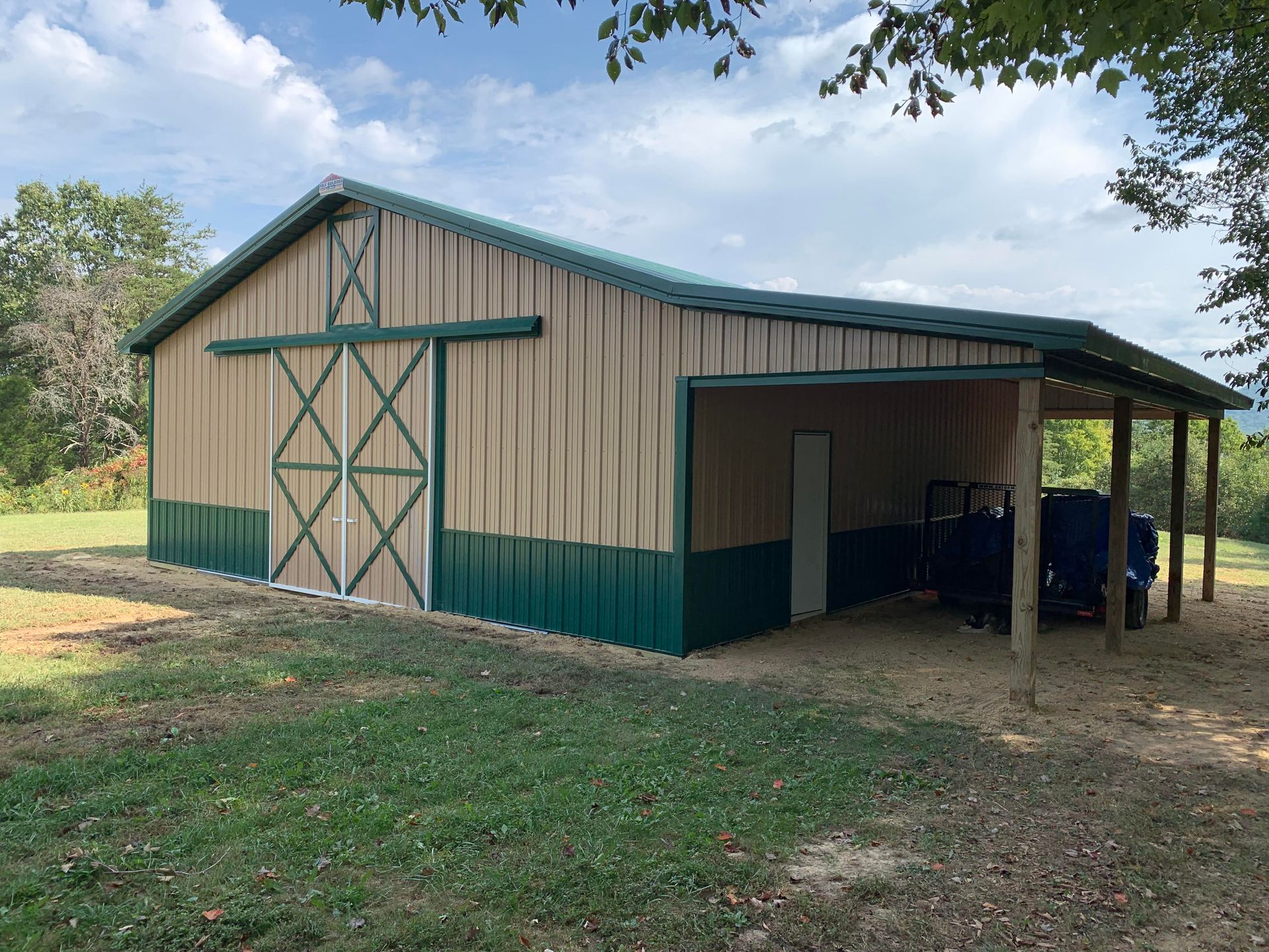 Tan and green metal barn with a carport, set in a grassy area under a partly cloudy sky.