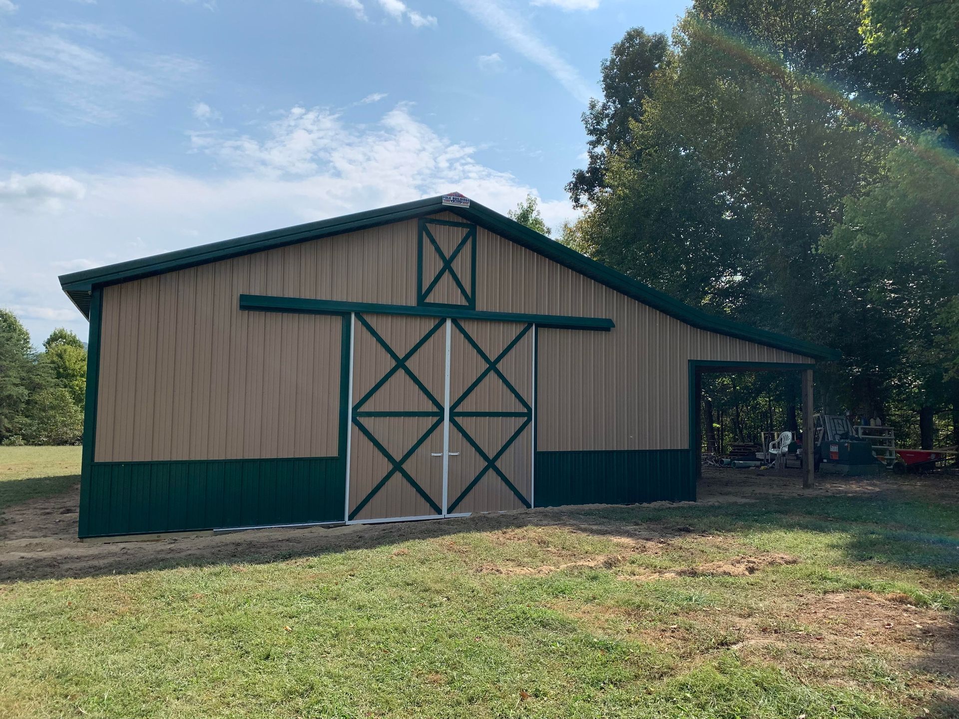 Tan and green barn with large sliding doors, set in a grassy yard with trees under a blue sky.