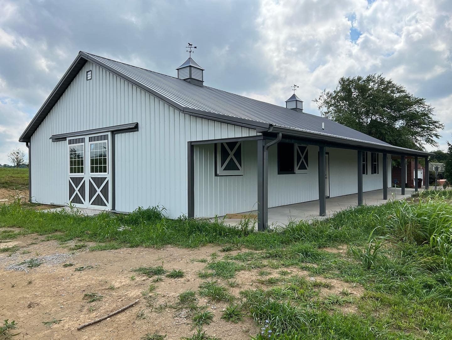White barn with a dark roof and porch, standing in a grassy area. The barn has a sliding door and several windows.