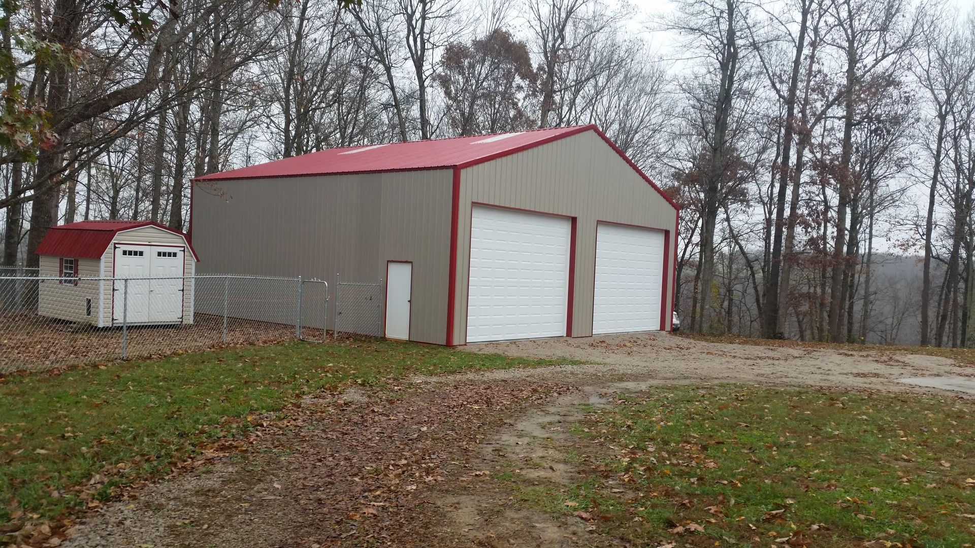A large tan garage with a red roof and two white garage doors sits on a grassy area with a smaller white shed to the left.