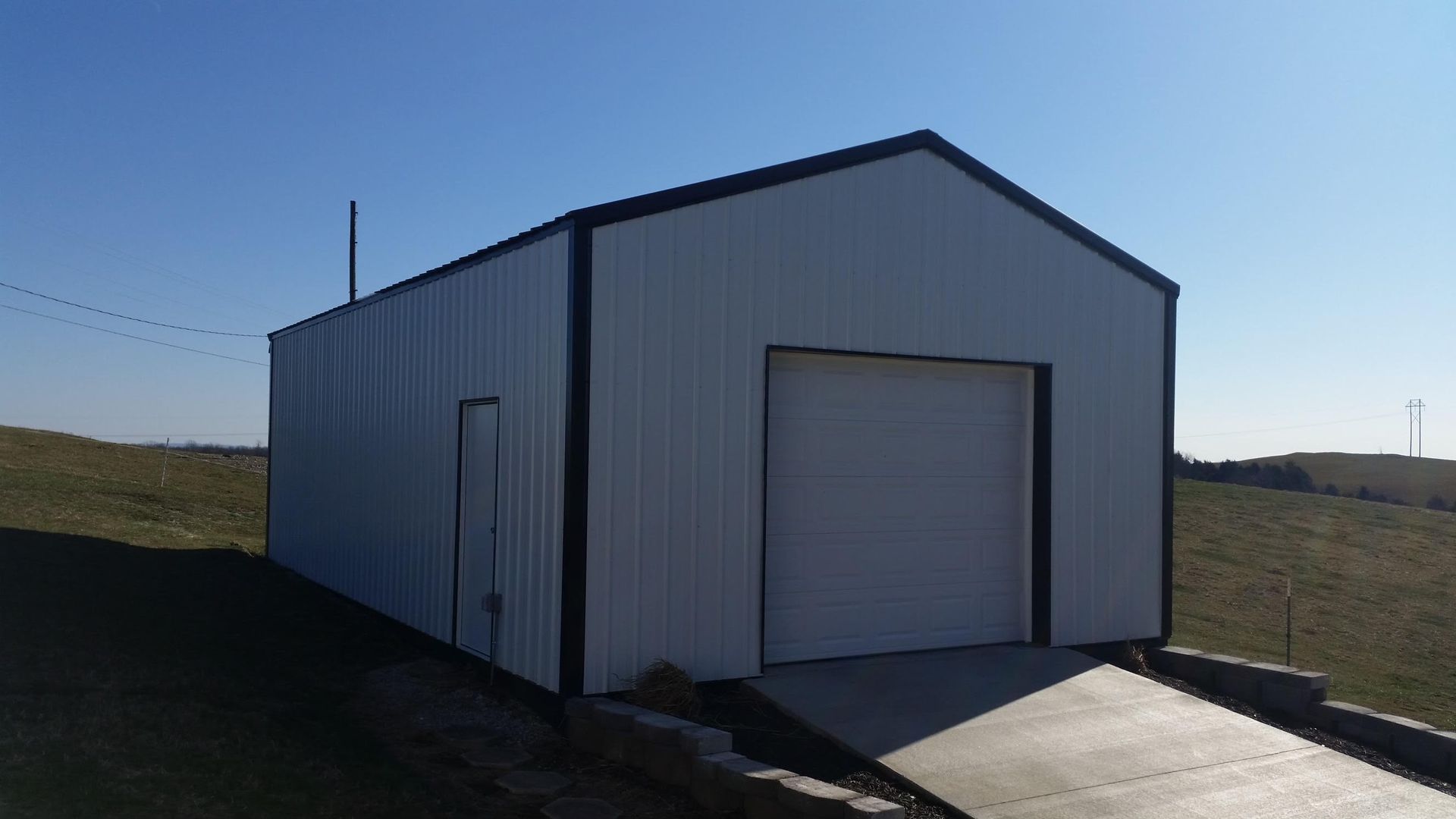 White metal storage building with a black trim, garage door, and entrance door, on a paved driveway on a grassy hill.