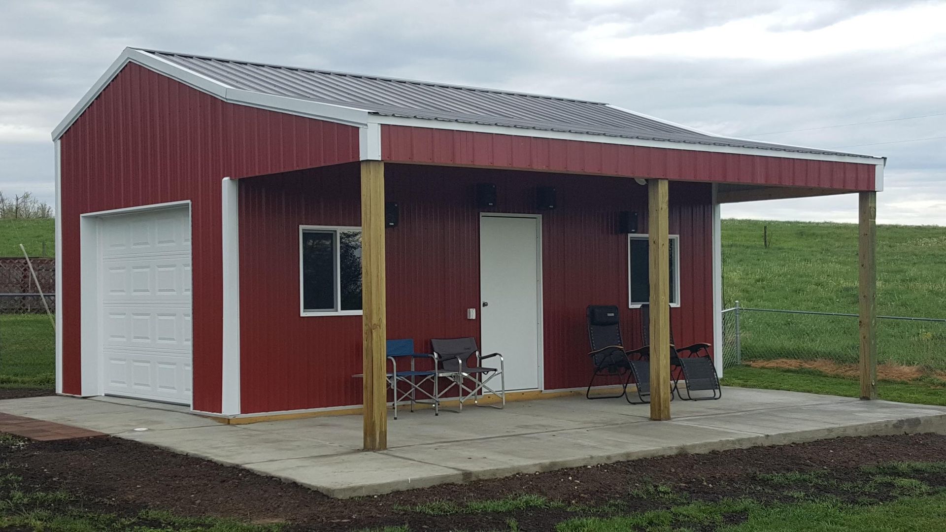 Red metal building with a porch, concrete foundation, and white garage door. Two windows and a white door are visible.