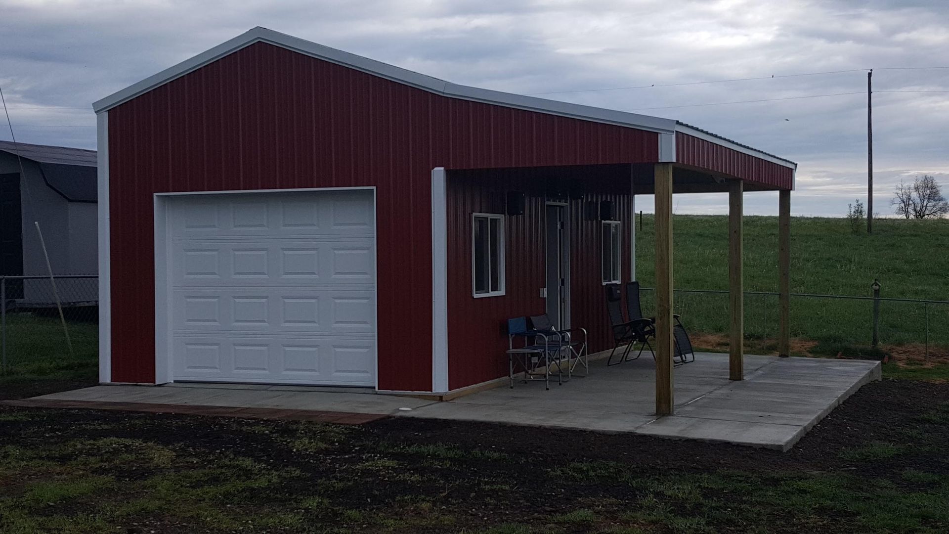 Red metal building with a white garage door and a covered porch on a concrete slab, set in a grassy field under a cloudy sky.