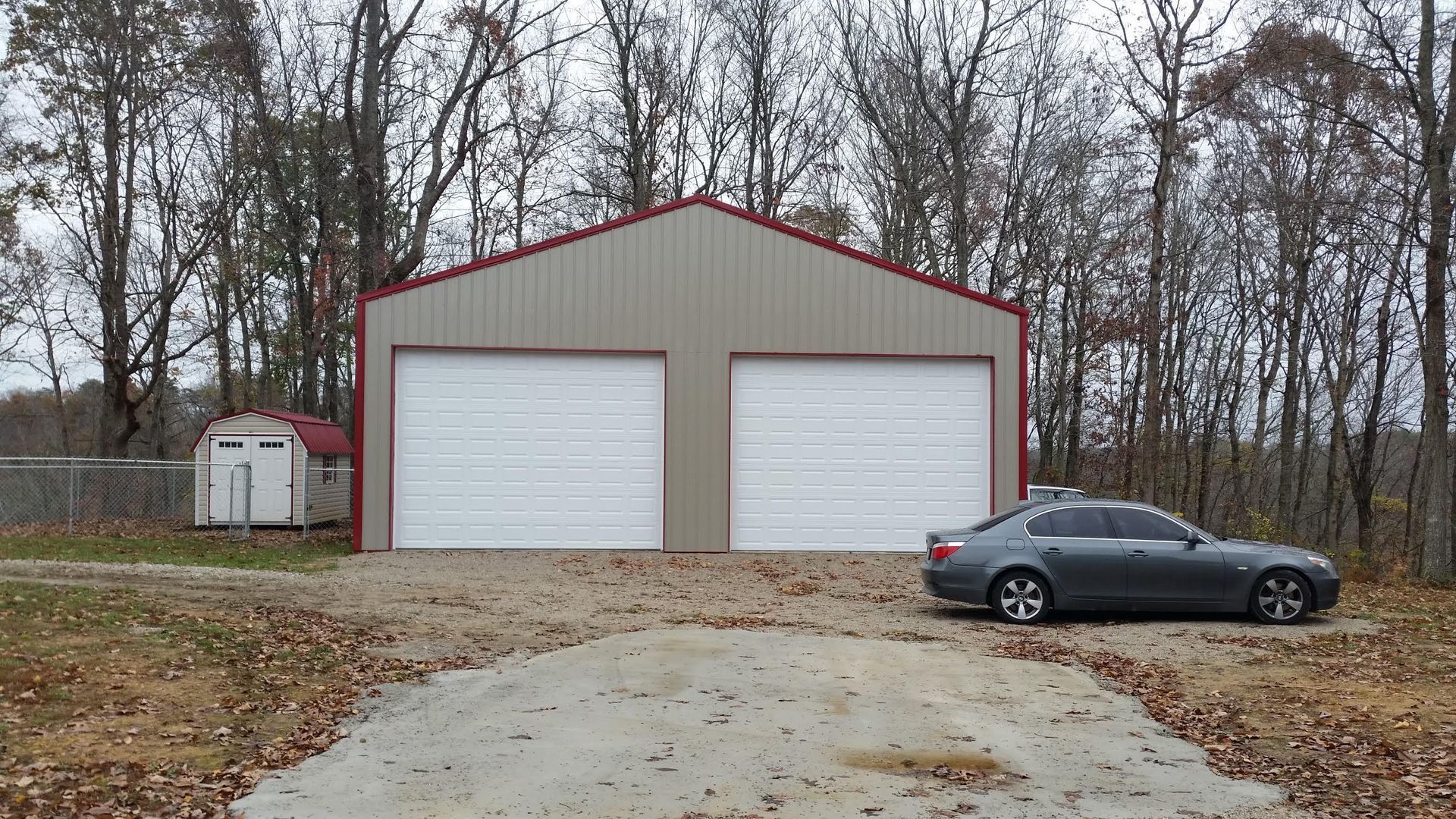 Two-car garage with red trim, gray siding, and two white garage doors. A gray car sits on the gravel driveway.