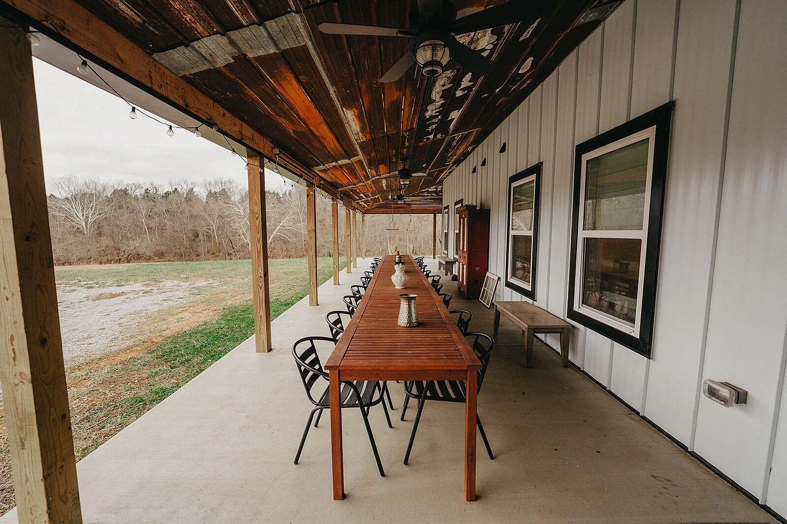 Covered porch with a long wooden table set for dining. Black chairs line the table, with windows and a bench against the white-sided building.