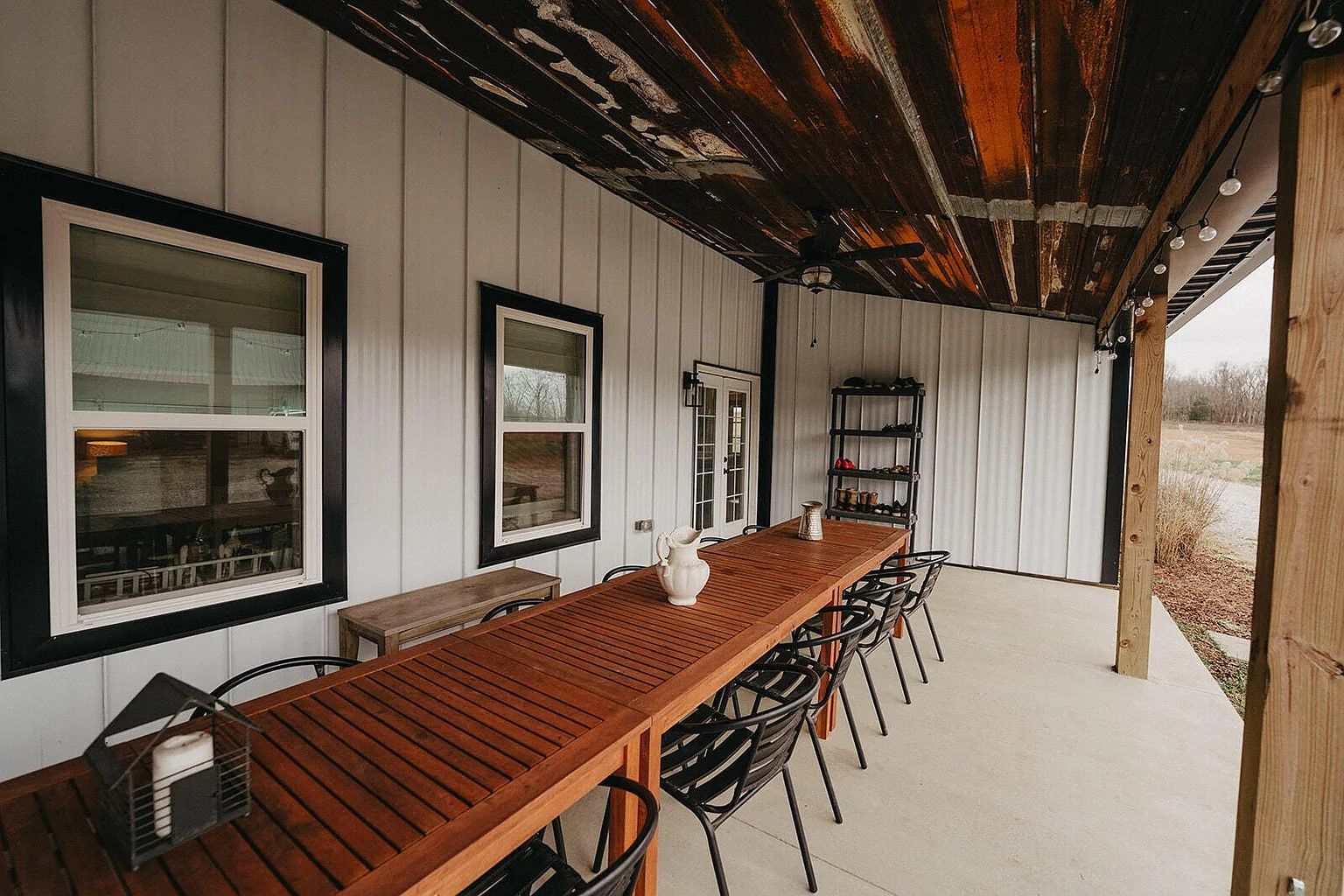 Covered outdoor dining area with a long wooden table and black chairs. White walls, dark window frames, and rustic ceiling.