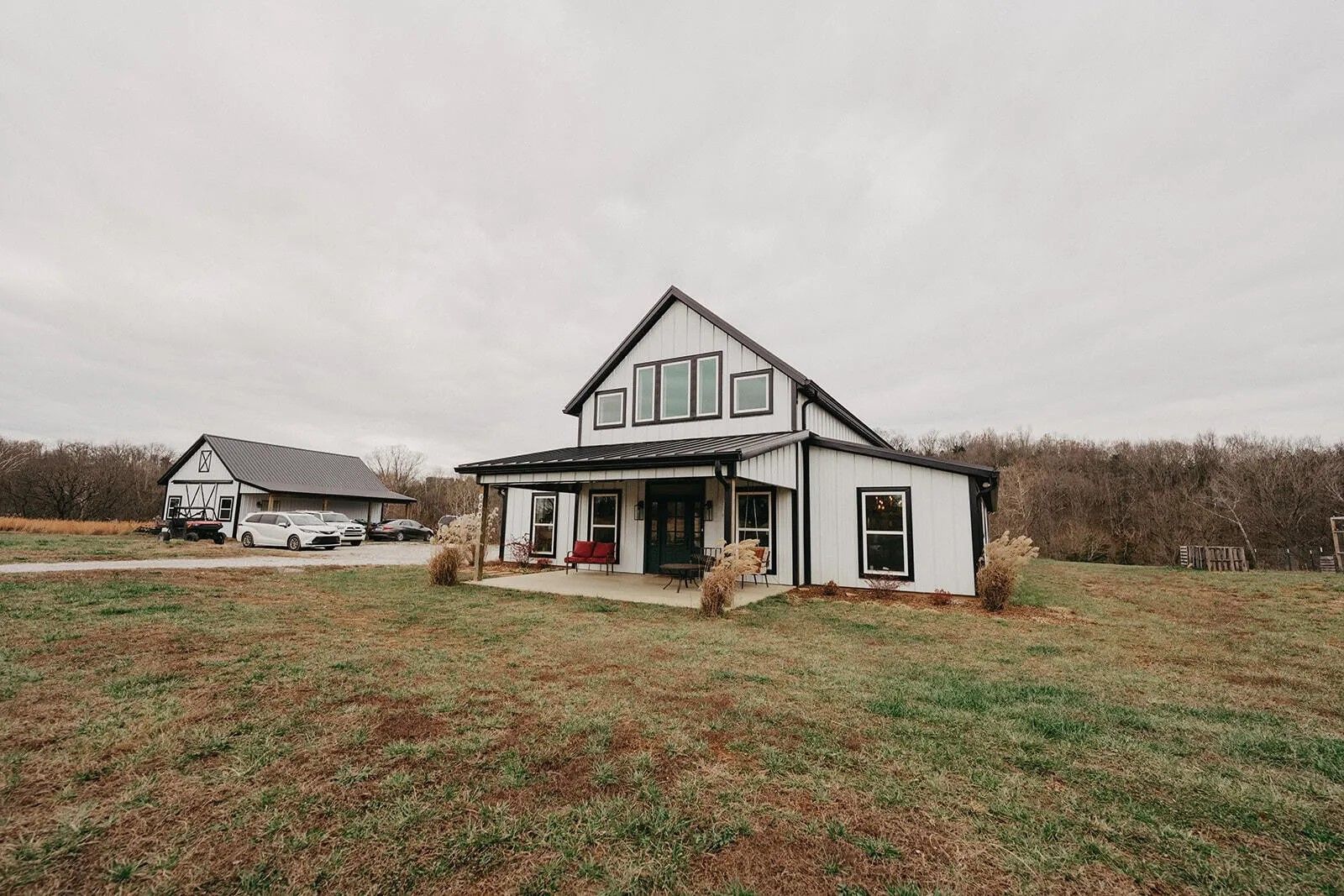 White farmhouse with black trim and a porch on a grassy field under an overcast sky; a smaller barn is in the background.