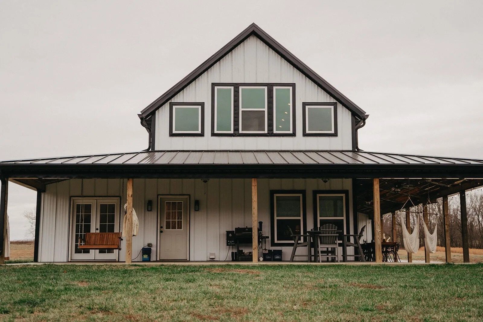 White farmhouse with a black roof and wrap-around porch on a cloudy day. The porch has a swing and seating.