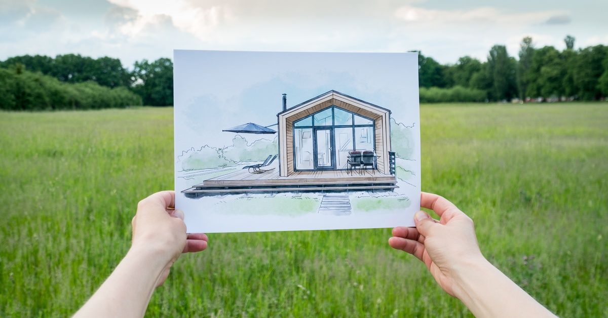 A person's outstretched hands hold a drawing of a pole barn house with a field, trees, and a partly 