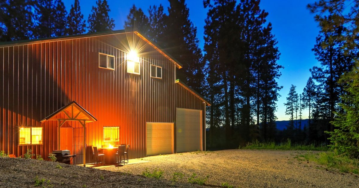 A large red metal barn home at night. It has two garage doors and a patio with a table, chairs, and a grill.