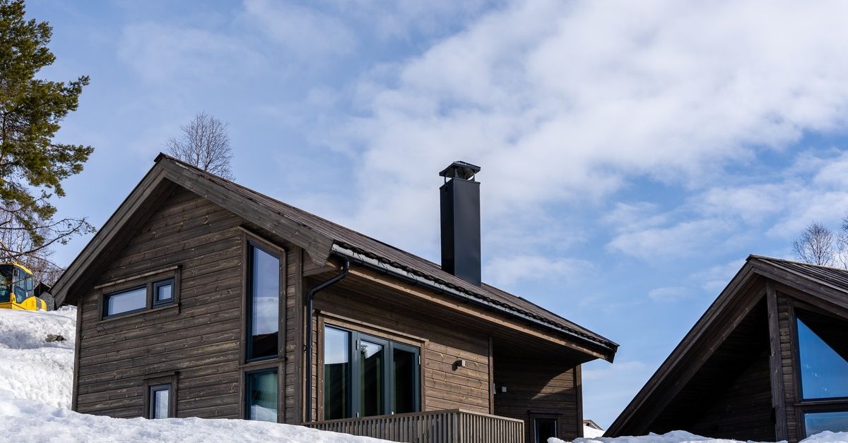 Two post-frame homes sit in a snowy landscape under a blue sky. They are wooden, and one has a porch and a chimney.