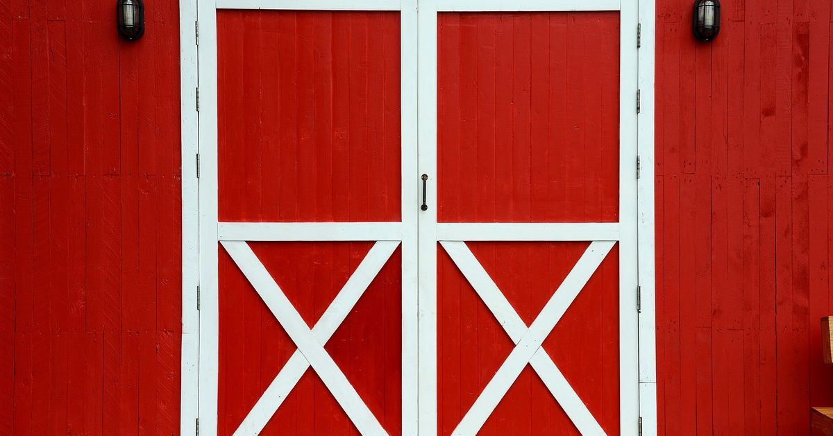 A close-up of a set of bright red barn doors with a classic white trim. There is an exterior light on either side of the doors.