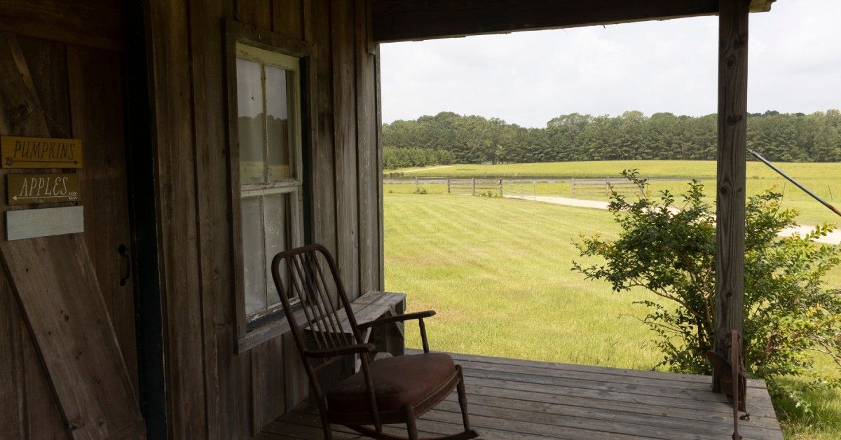 A wooden porch attached to a home. A large field stretches beyond the porch, and a decorative sign reads,