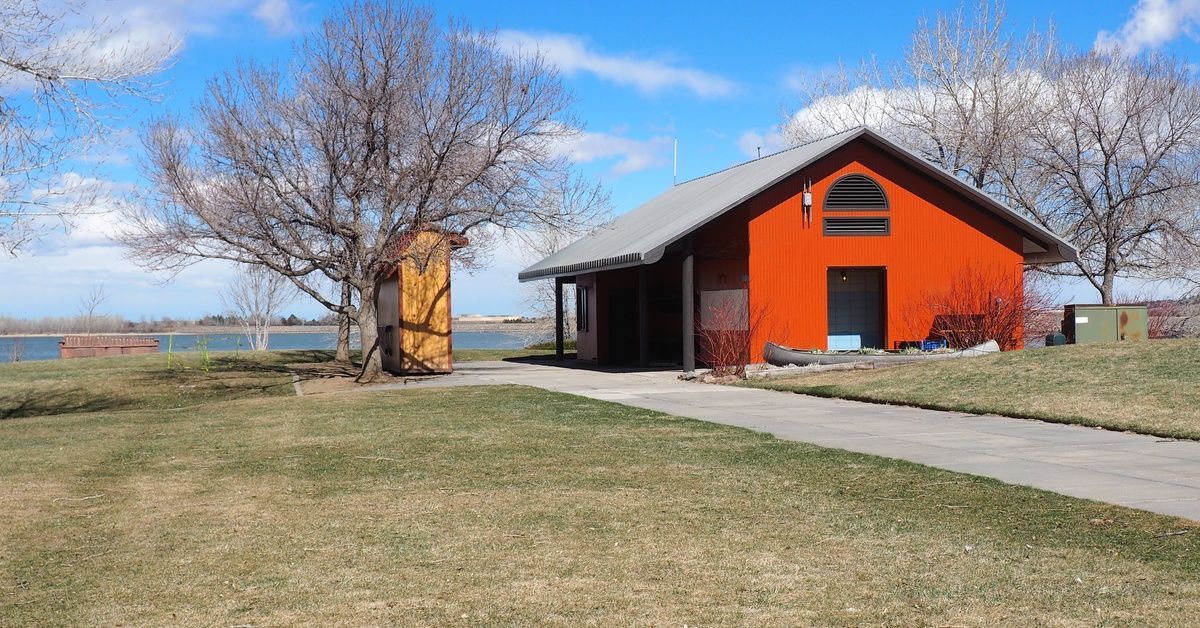 A bright red pole barn with a gray roof sits near the edge of a body of water with a sidewalk leading to it.
