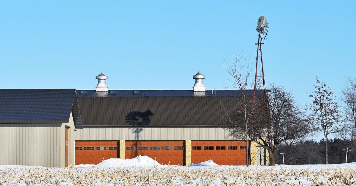 A large barn with three garage doors sits next to a windmill in a snowy field with bare trees agains