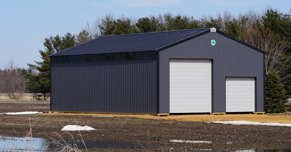 A dark gray pole building with two white garage doors. Patches of snow are melting around it, and trees stand behind it.
